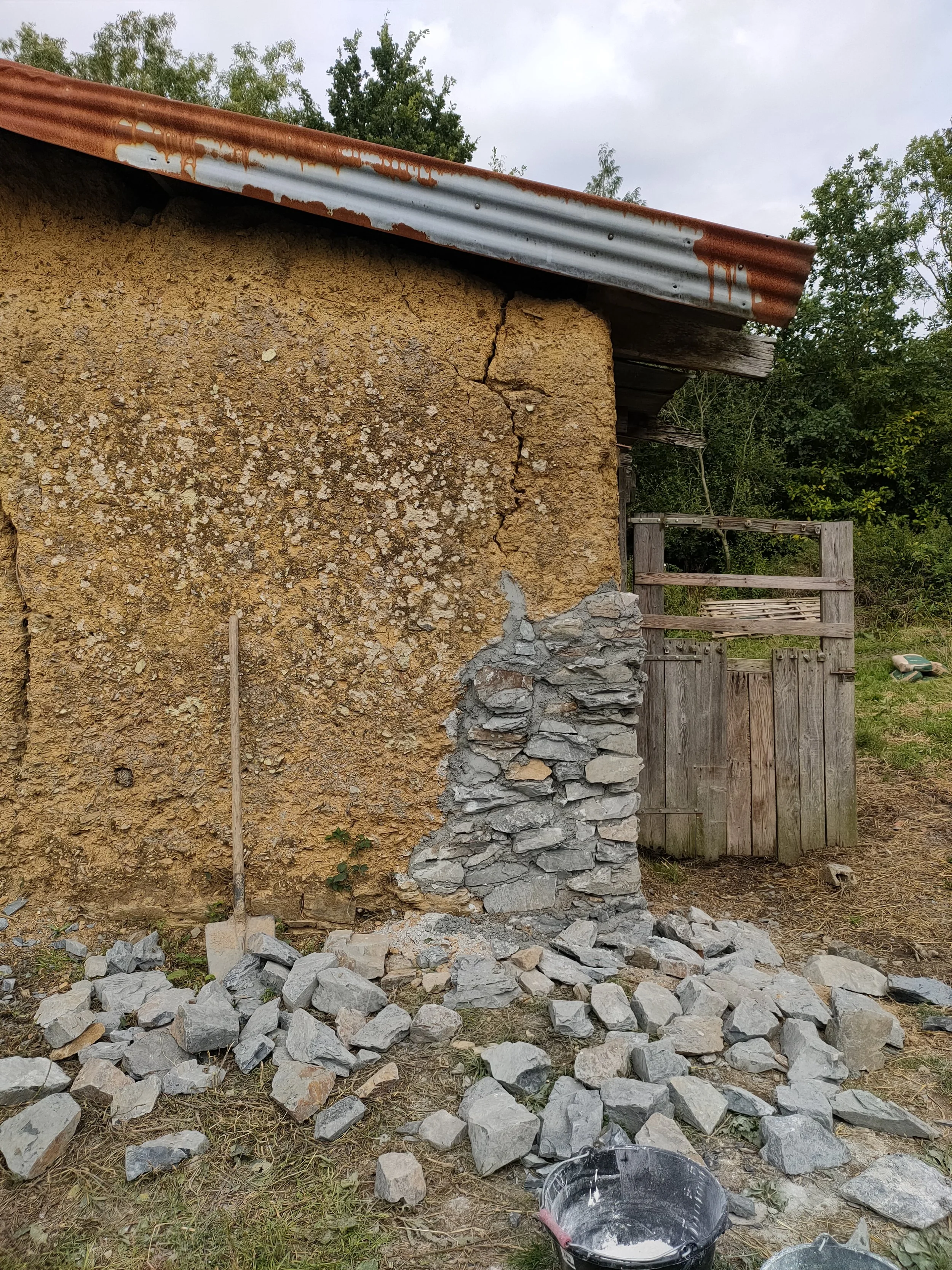Une vieille maison en terre et pierres avec une fissure sur le mur extérieur, un toit en tôle rouillée, une porte en bois, et des pierres empilées pour réparer la fissure. Un seau avec du mortier de chaux est au sol.