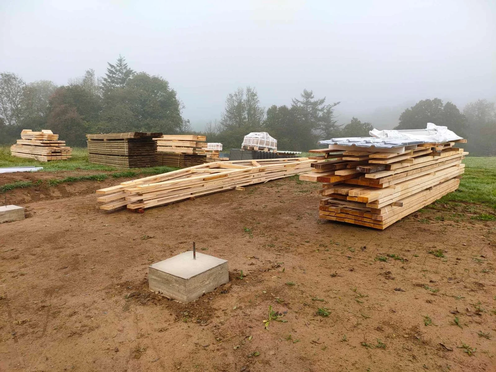 Tas de planches de bois empilées sur un terrain de construction, avec un fond de bocage et un ciel brumeux.