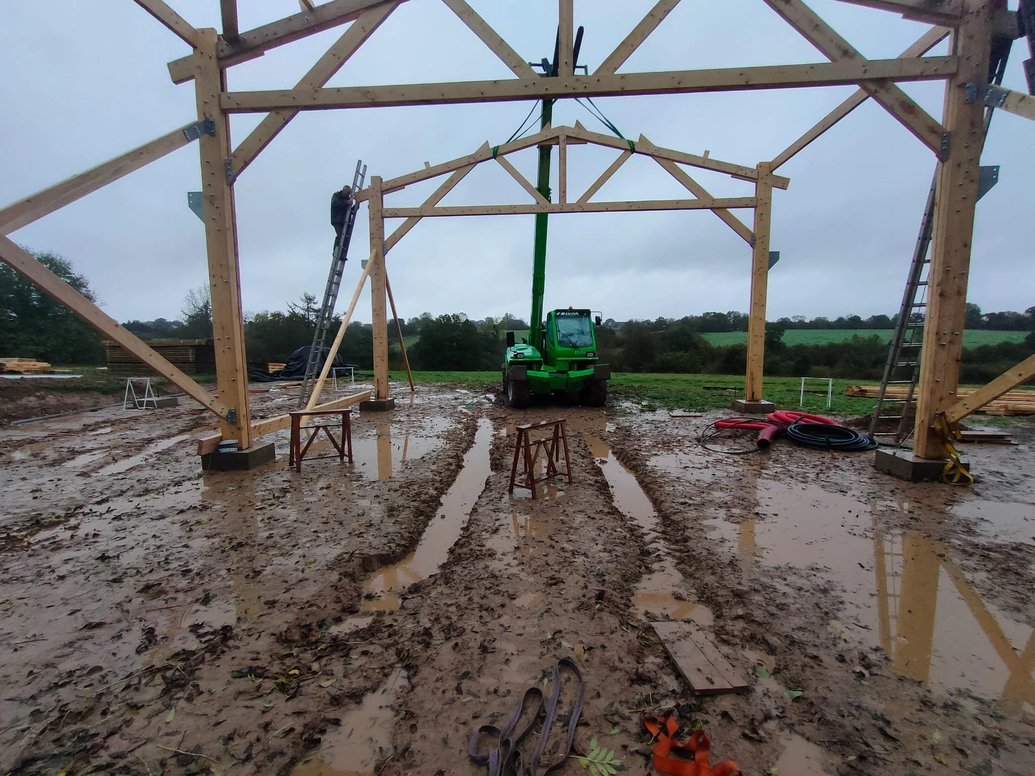 Construction d'une structure en bois sous un ciel nuageux, avec un sol boueux et des équipements comme des échelles et une machine verte de levage.