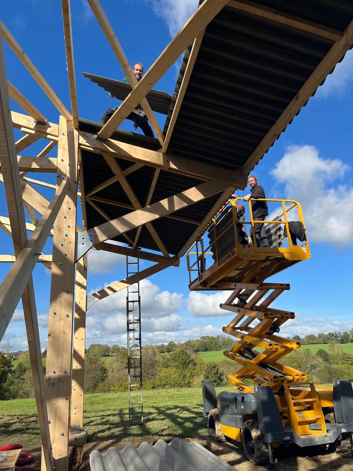 Deux hommes couvrent une charpente en bois avec une plateforme élévatrice sous un ciel bleu avec quelques nuages.