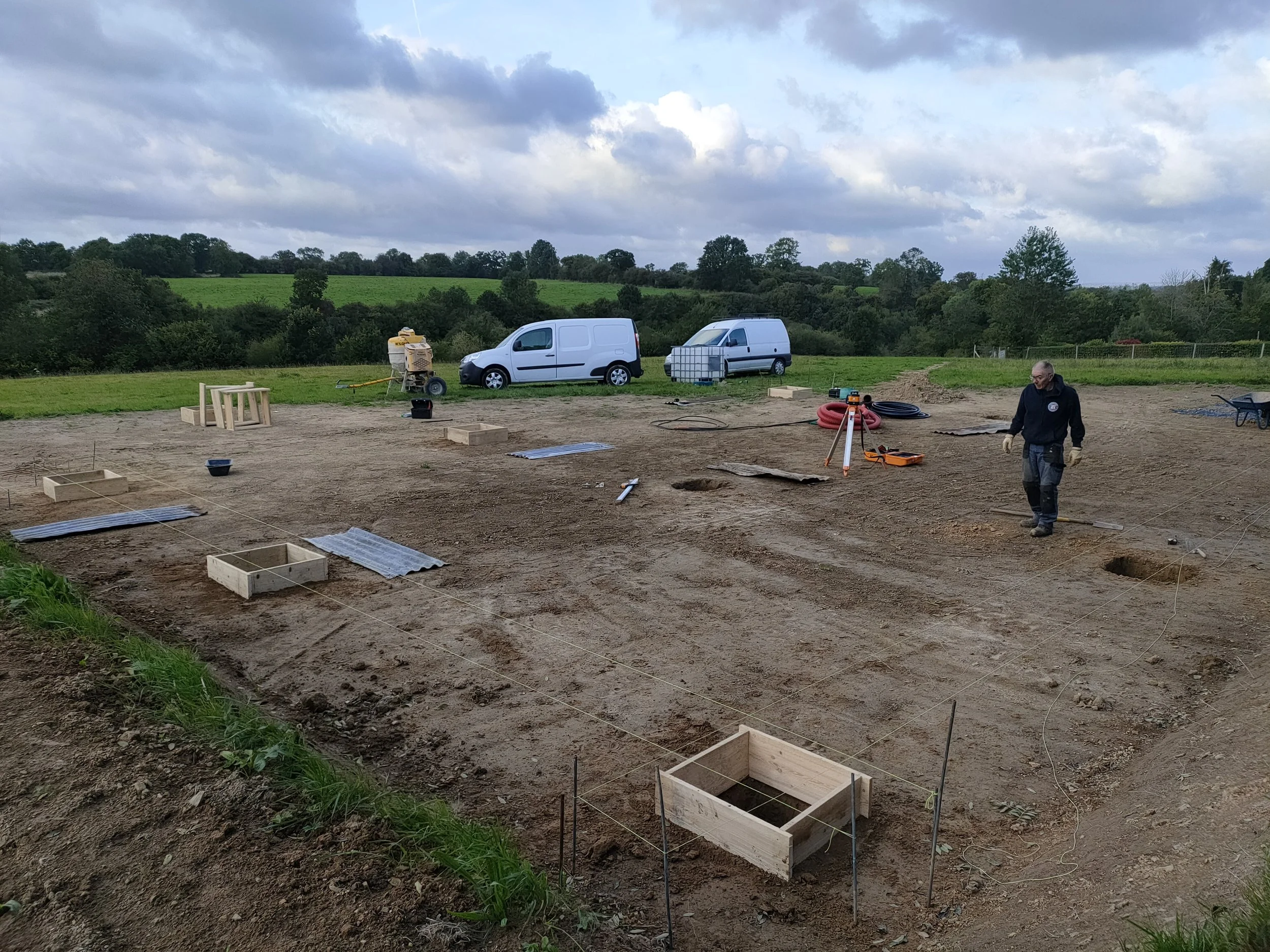 Chantier de construction en plein air avec un homme portant des équipements de sécurité, des cadres en bois, des outils, deux véhicules blancs en arrière-plan, et un paysage rural avec des arbres et un ciel nuageux.