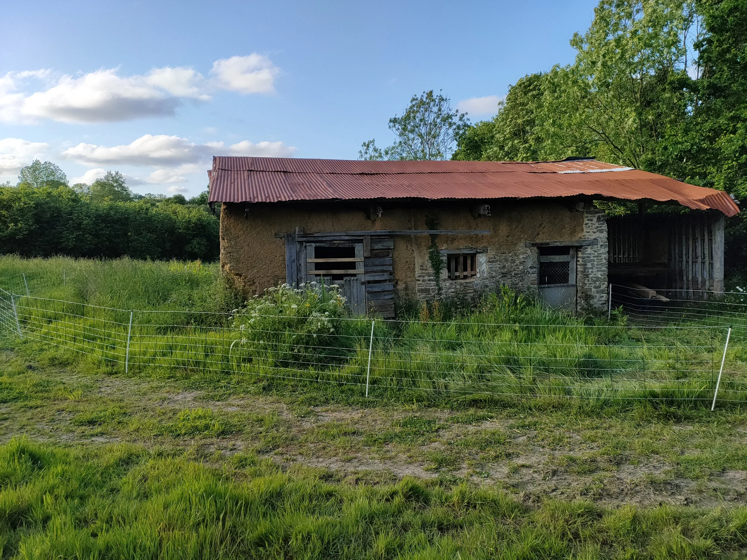 Un vieux bâtiment en pierre et en terre avec un toit en tôle rouillée, entourée d'herbe verte et d'une clôture en fil métallique dans un environnement rural, sous un ciel bleu avec quelques nuages.