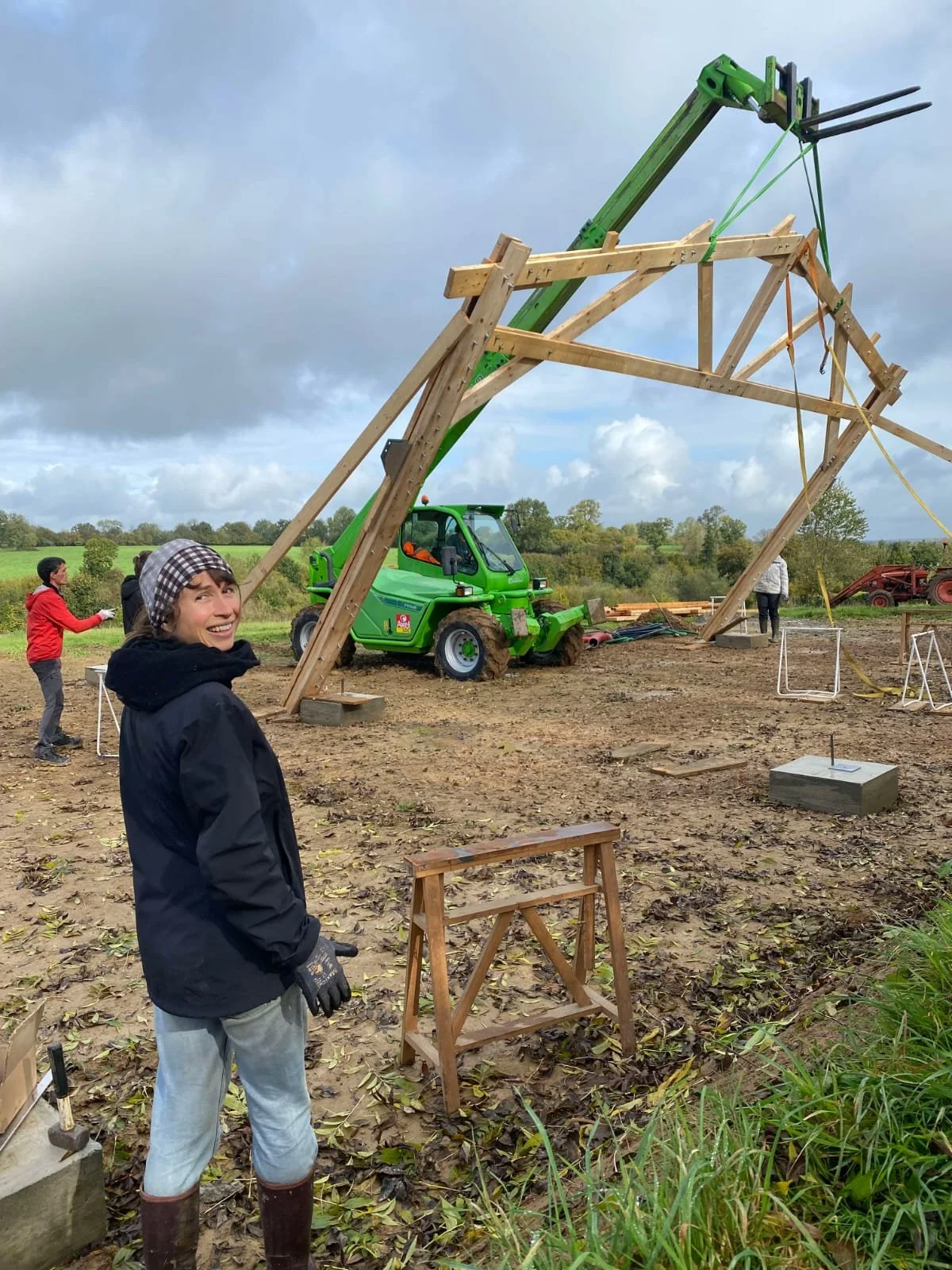 Une femme souriante en tenue de travail se tient devant une structure en bois en construction sur un terrain boueux, avec un téléscopique vert à proximité et d'autres personnes en arrière-plan.