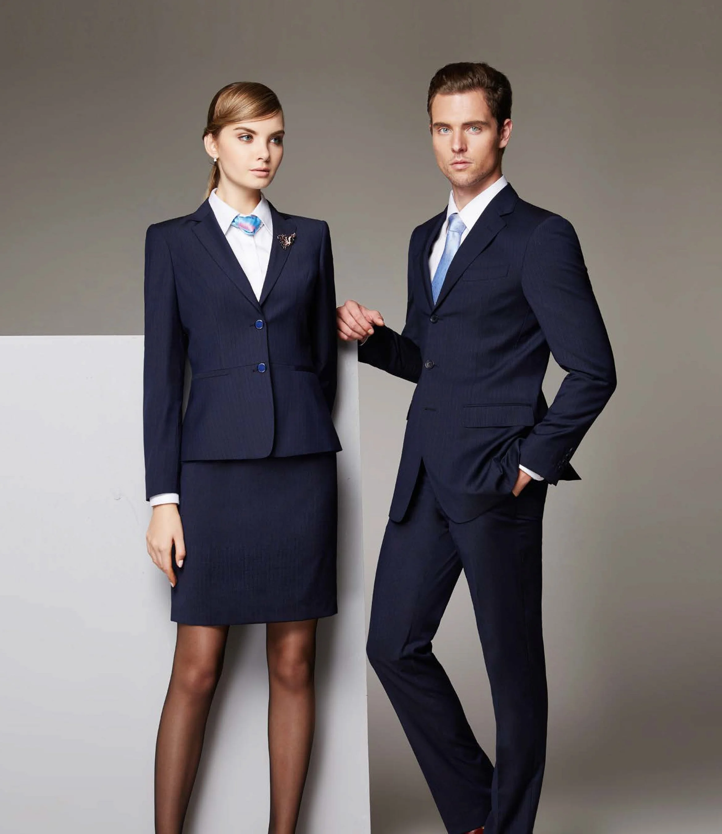 A man and woman dressed in dark business suits standing against a neutral background. The woman is wearing a blazer and skirt, and the man is wearing a suit with his hand in his pocket.