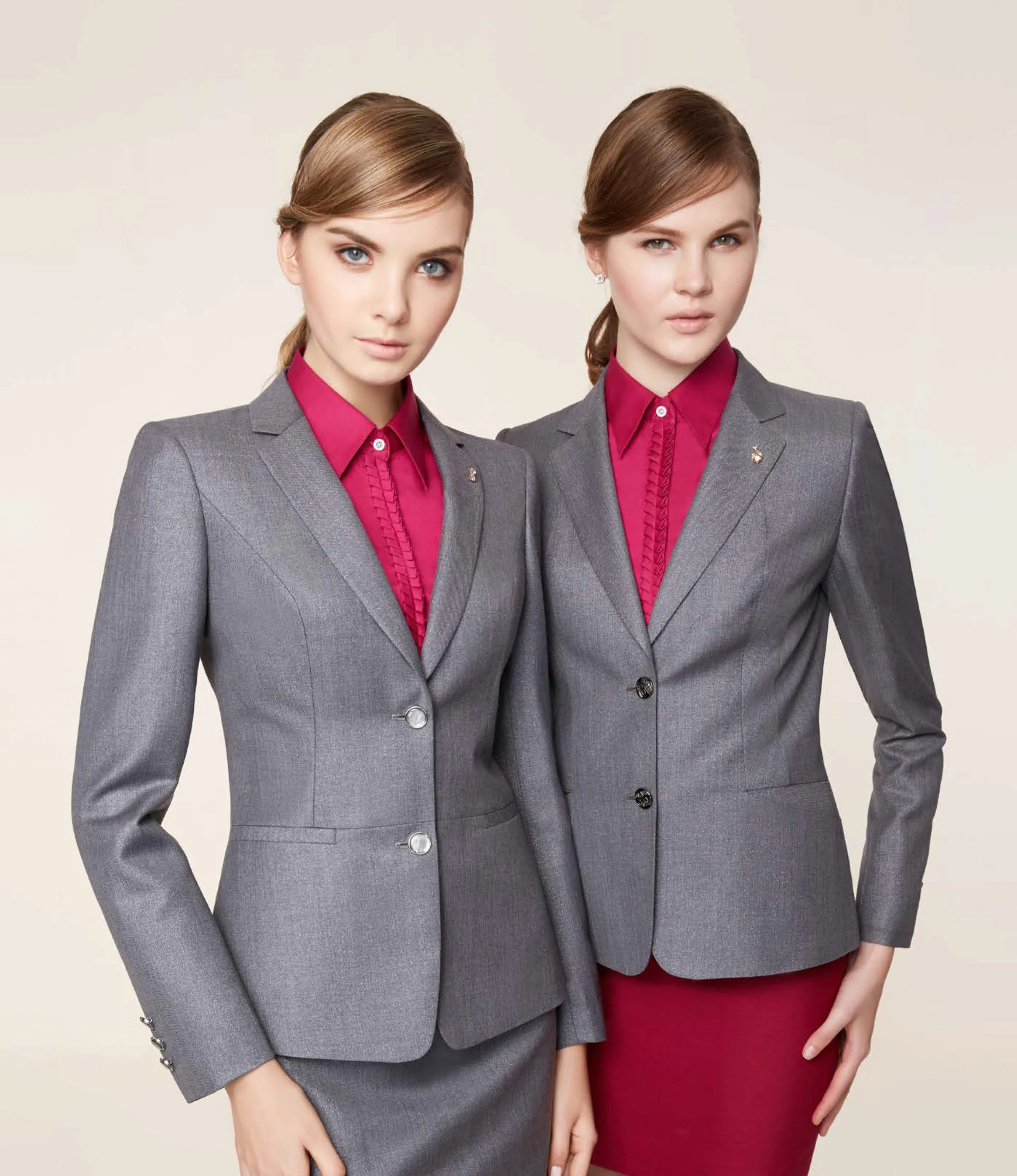 Two young women wearing gray business suits with pink blouses standing against a plain background.