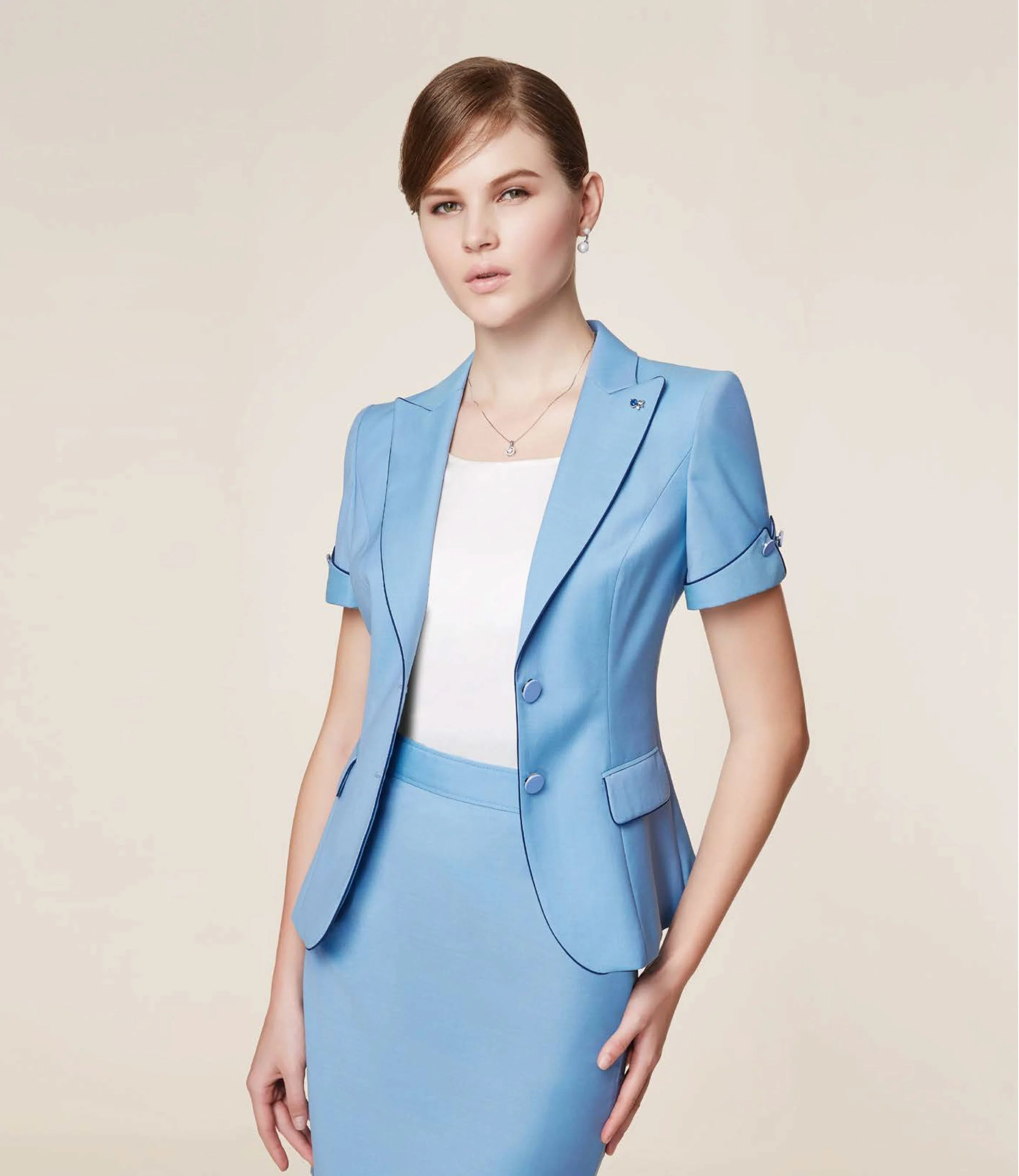 A woman wearing a light blue blazer and matching skirt, paired with a white top, standing against a neutral background.