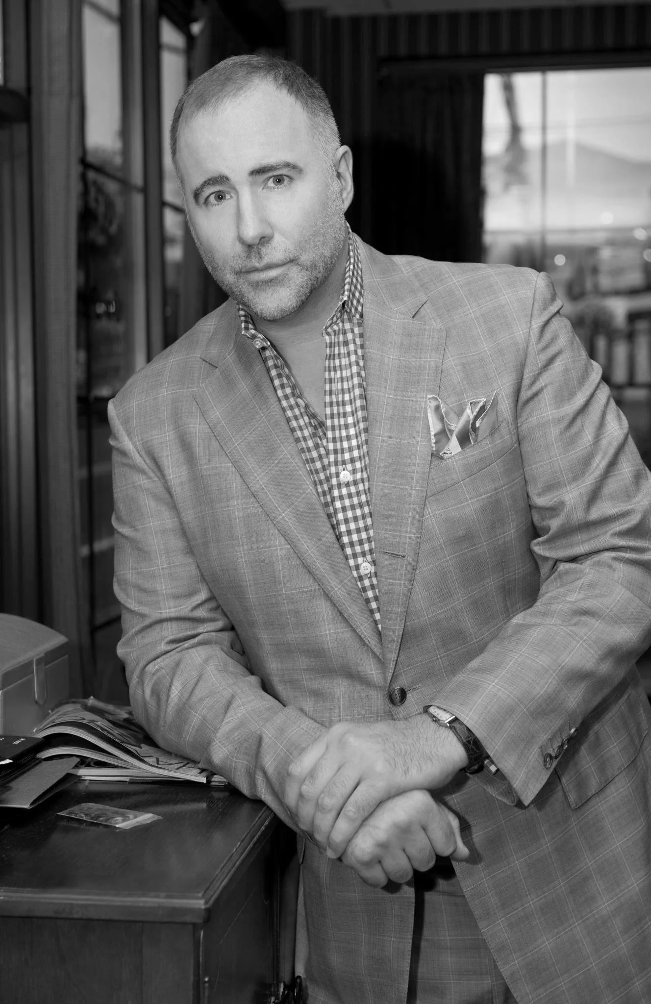Black and white photo of a man in a plaid suit jacket and checkered shirt, standing with arms crossed at a counter in a cafe or restaurant.