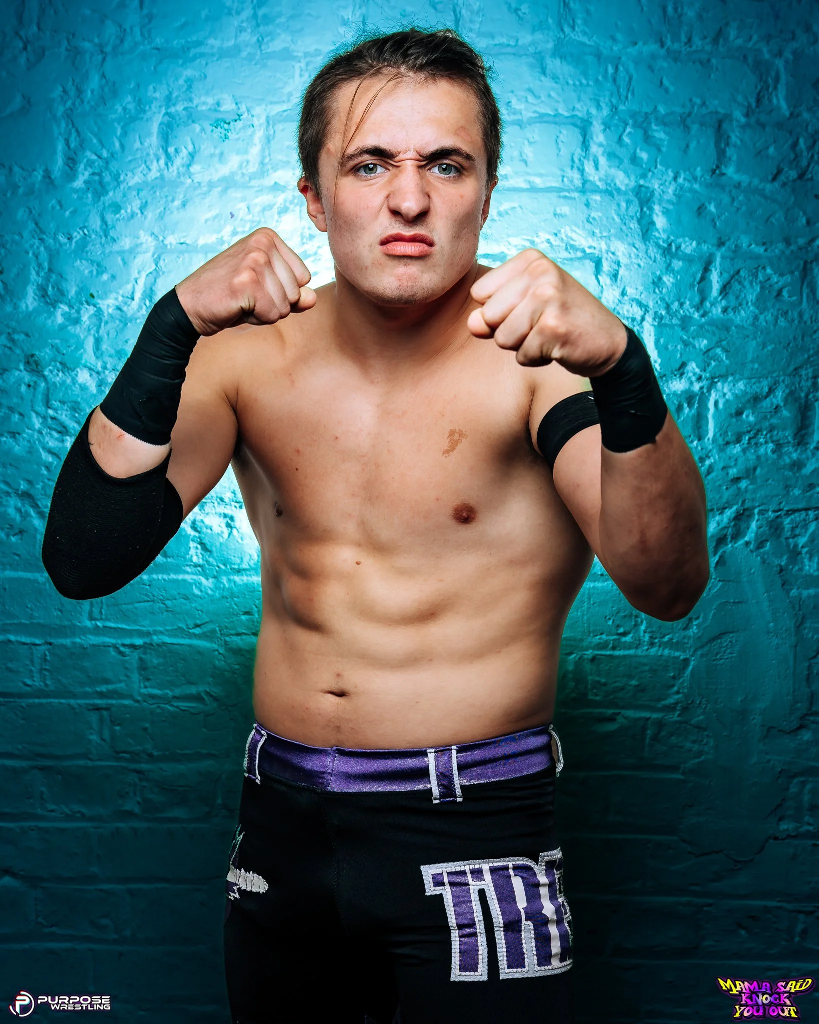 Young male wrestler with black wrist wraps posing shirtless in front of a blue brick wall, with a determined expression and clenched fists.