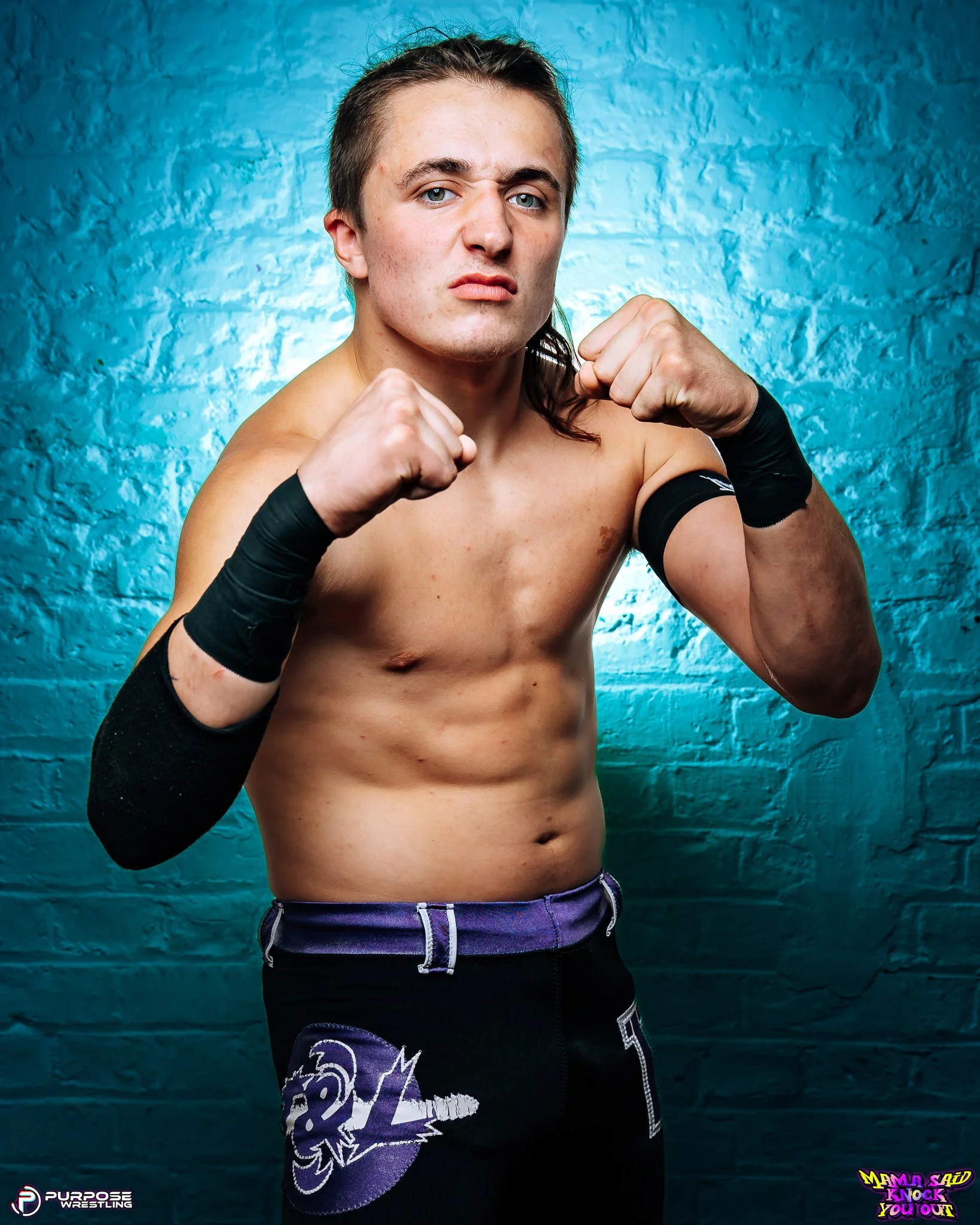 A young male wrestler with tan skin and long hair tied back, shirtless with black wrist guards, posing with fists up in a fighting stance against a blue brick wall background.