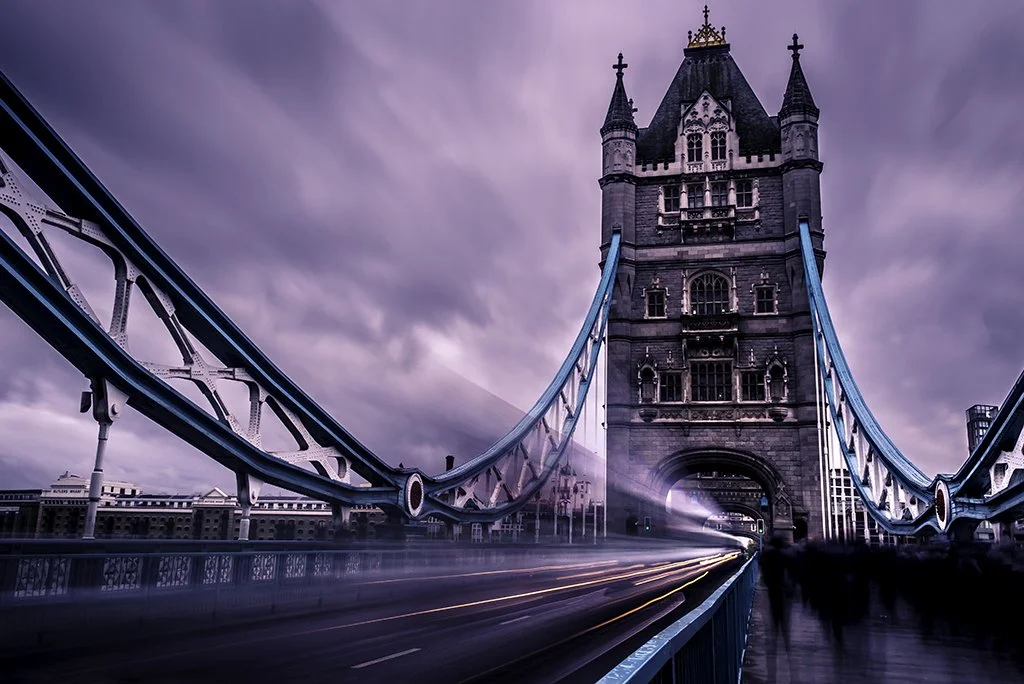 The Tower Bridge in London with a cloudy sky, illuminated suspension cables, and blurred moving vehicles and pedestrians on the bridge.
