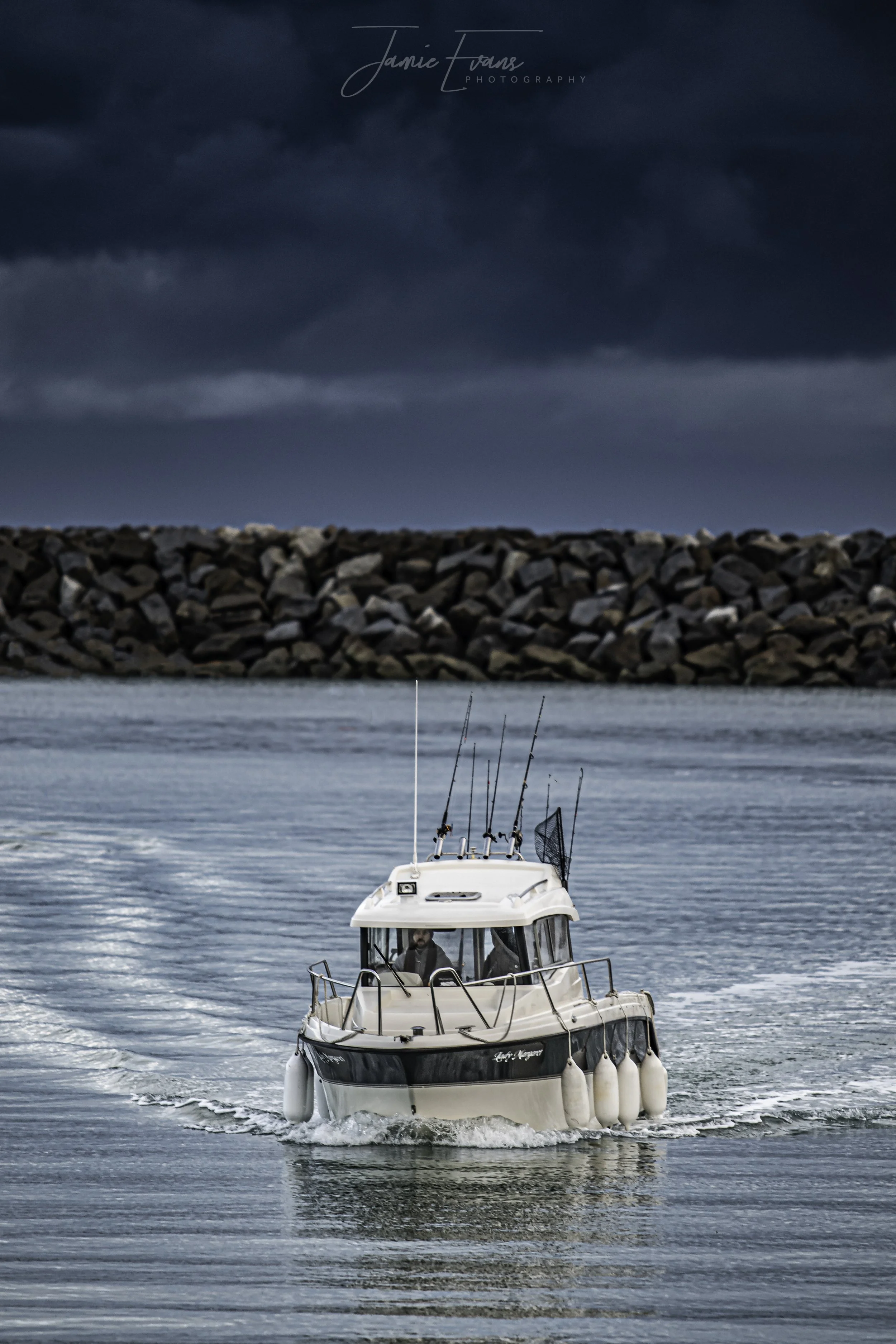 A boat on calm water with fishing poles, approaching a rocky breakwater under a stormy sky.