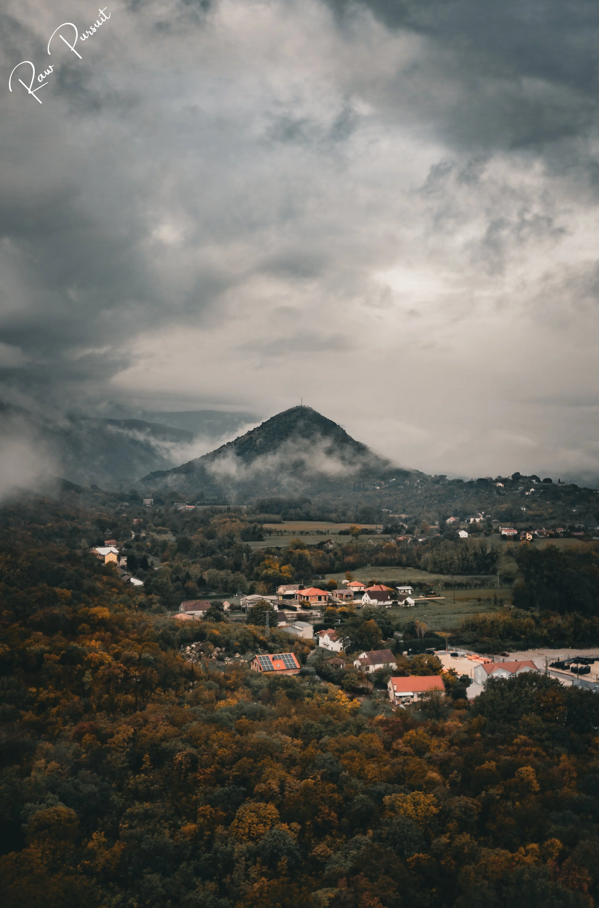 A scenic view of a mountain surrounded by clouds and a small town nestled in a forest below, with a cloudy sky overhead.