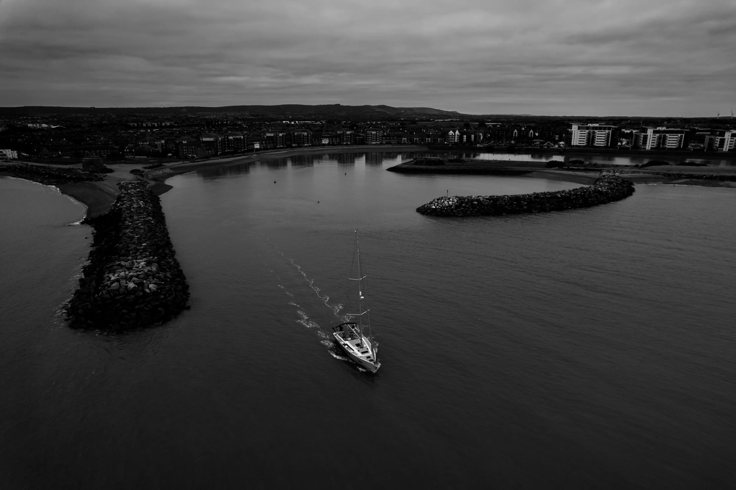 A black and white aerial view of a boat sailing through a harbor with breakwaters, a city skyline, and hills in the background.