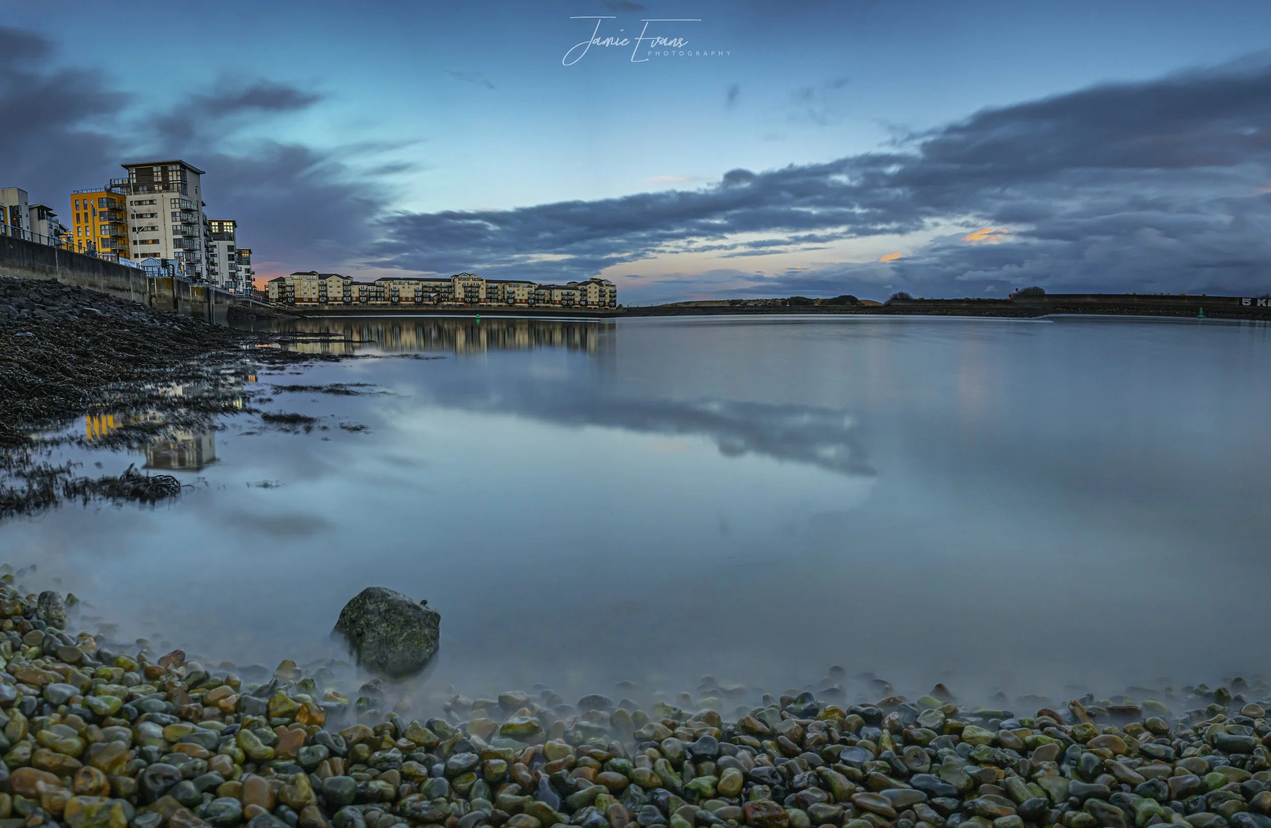Night view of a calm body of water reflecting the sky and buildings, with a pebble beach and rocks in the foreground and a cityscape of apartment buildings on the left side.
