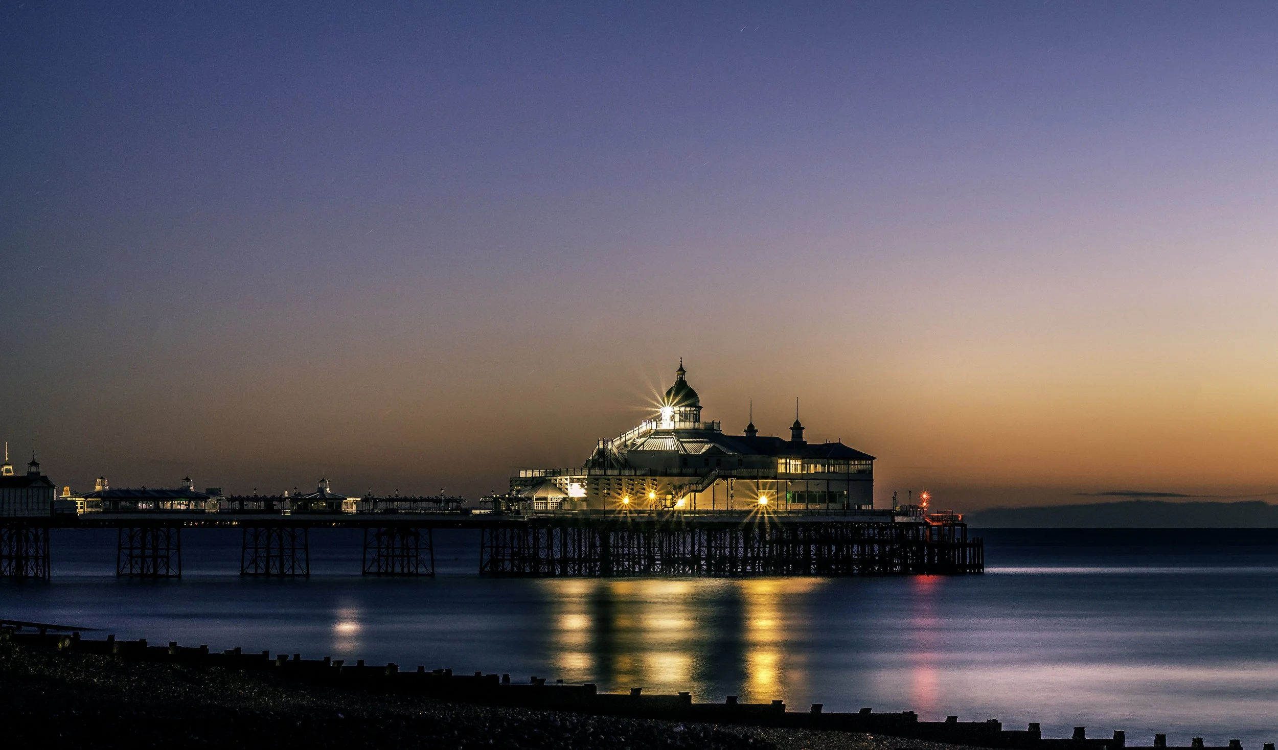 Sunset over a pier with a lit-up building on the water.