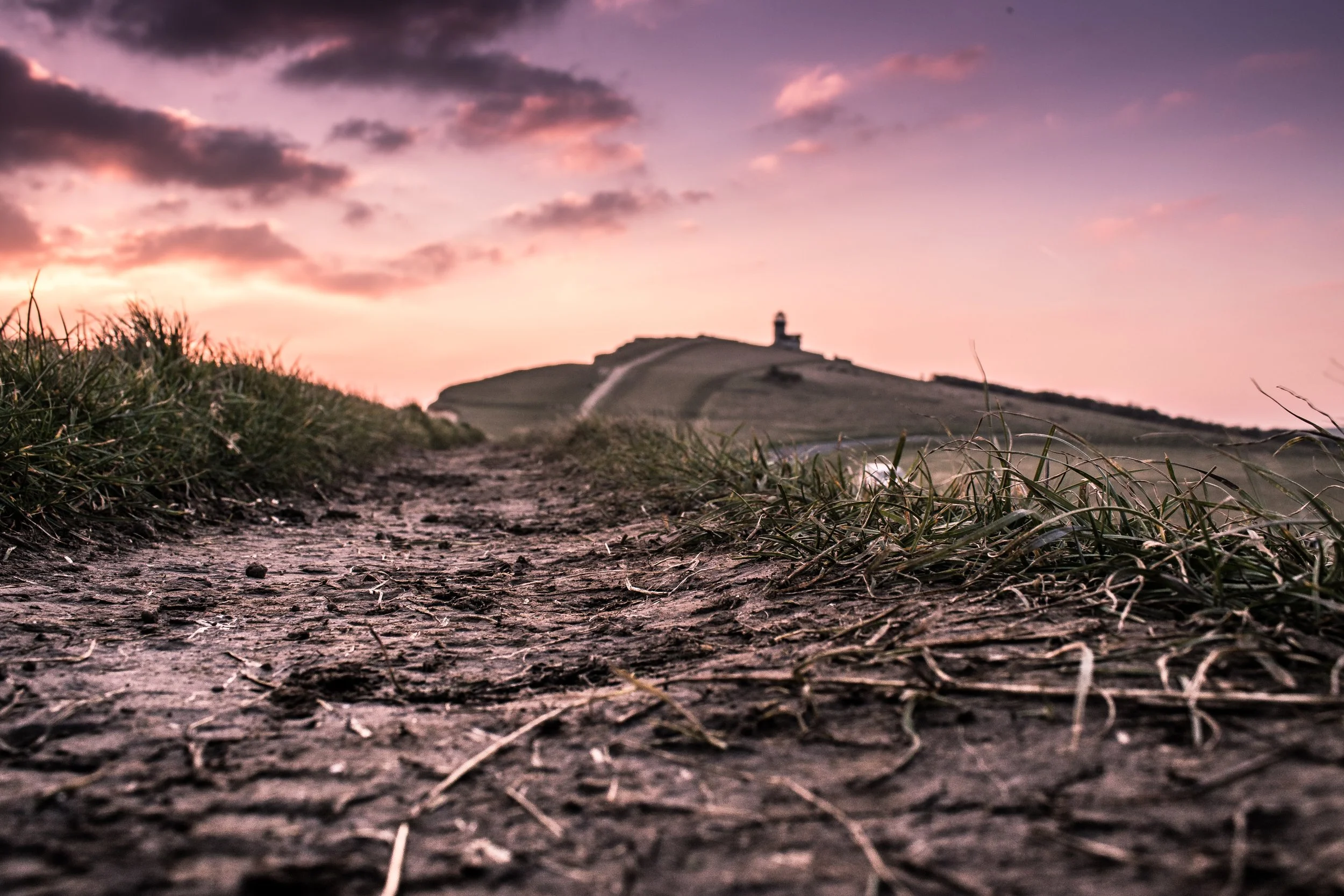 A dirt path leading towards a hill with a lighthouse on top during sunset, with pink and purple clouds in the sky.