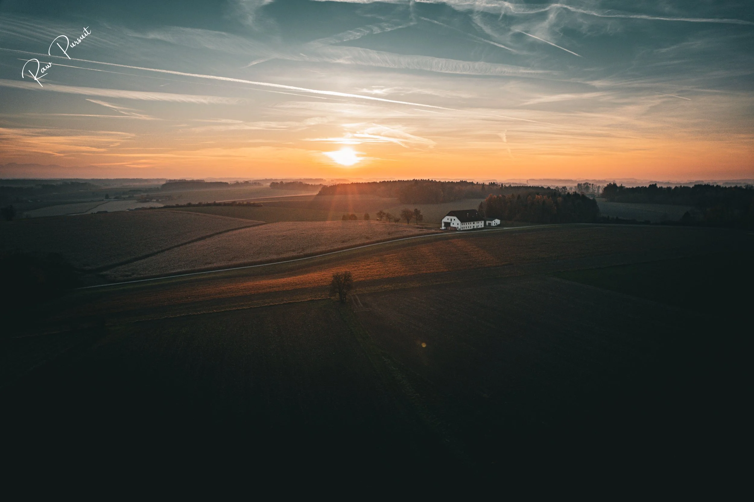 A scenic view of a rural landscape at sunset, featuring a white house with a black roof, rolling fields, and a winding road. The sky is partly cloudy with streaks and contrails, and the sun is near the horizon casting a warm glow.