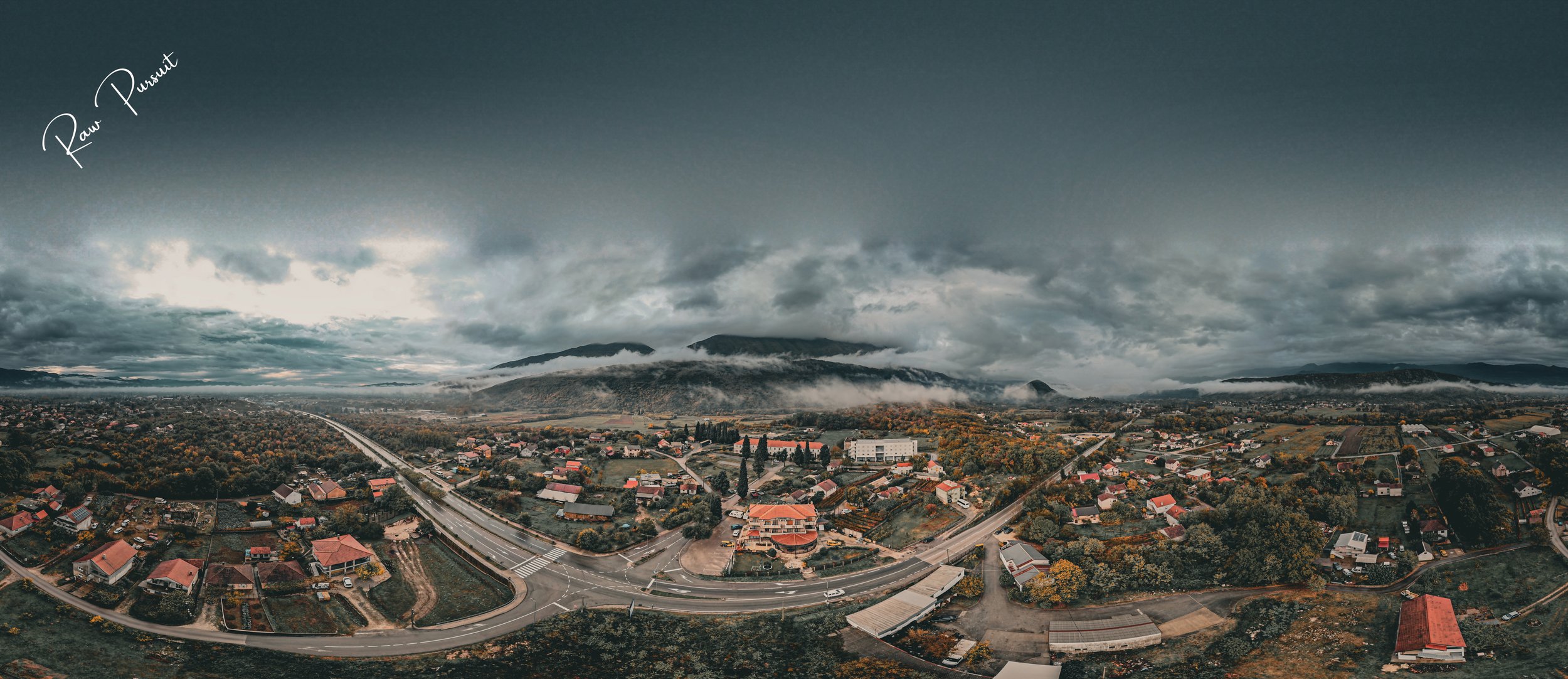 Panoramic view of a small town with houses, roads, and surrounding lush green trees under a cloudy sky with mountains in the background.