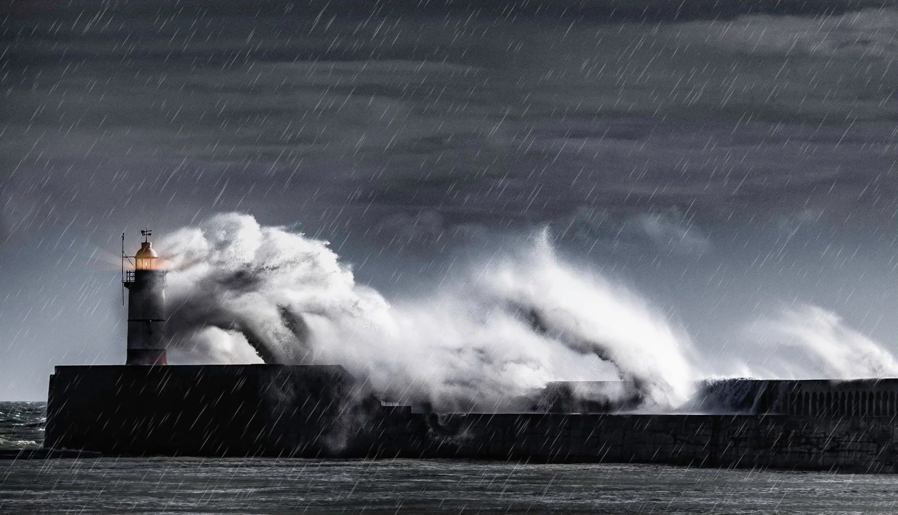 A lighthouse on a pier being battered by stormy seas with large, crashing waves and rain during a storm.