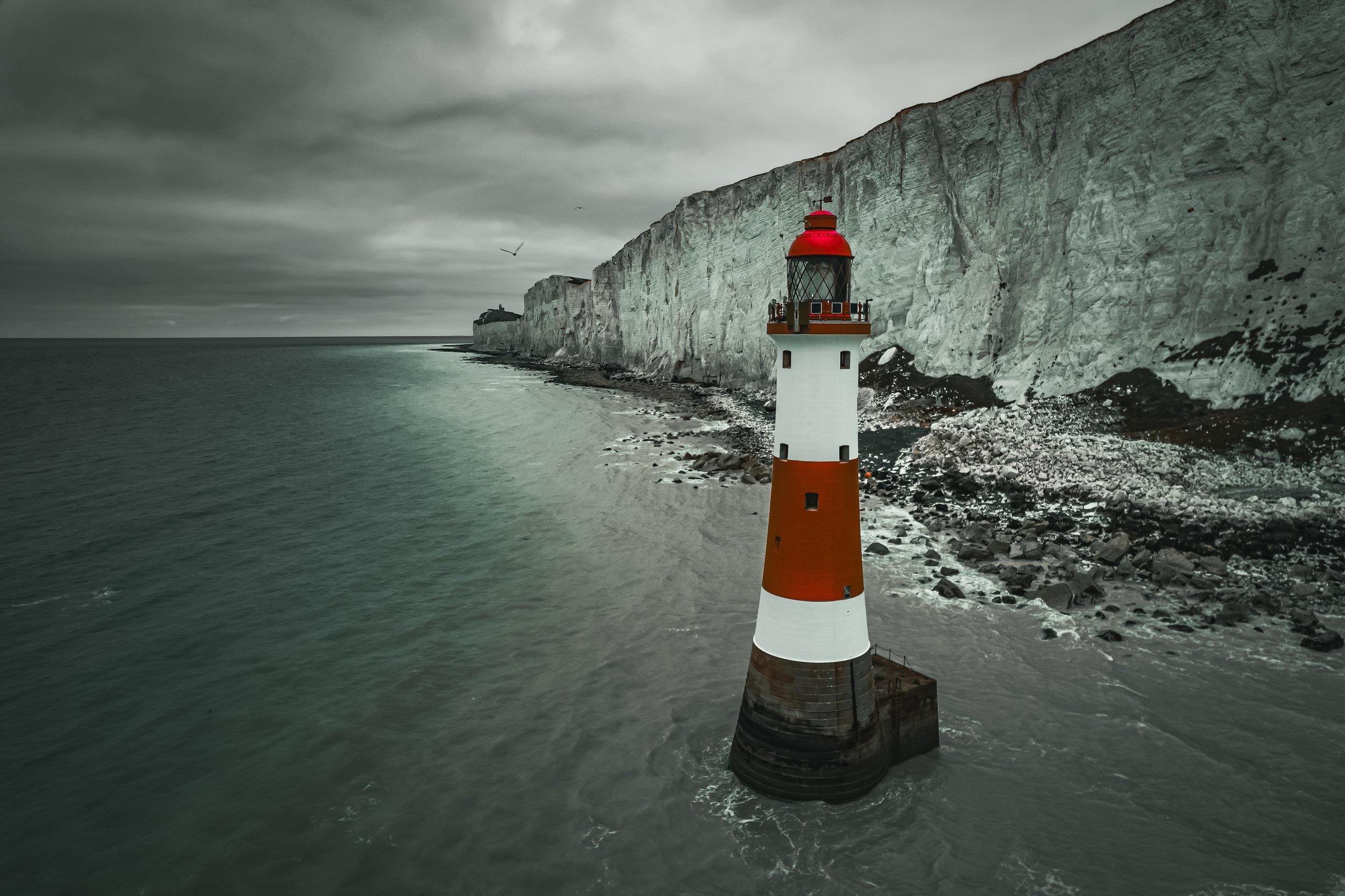 A lighthouse with a red top and white and orange striped body standing in the water near white chalk cliffs on a cloudy day.