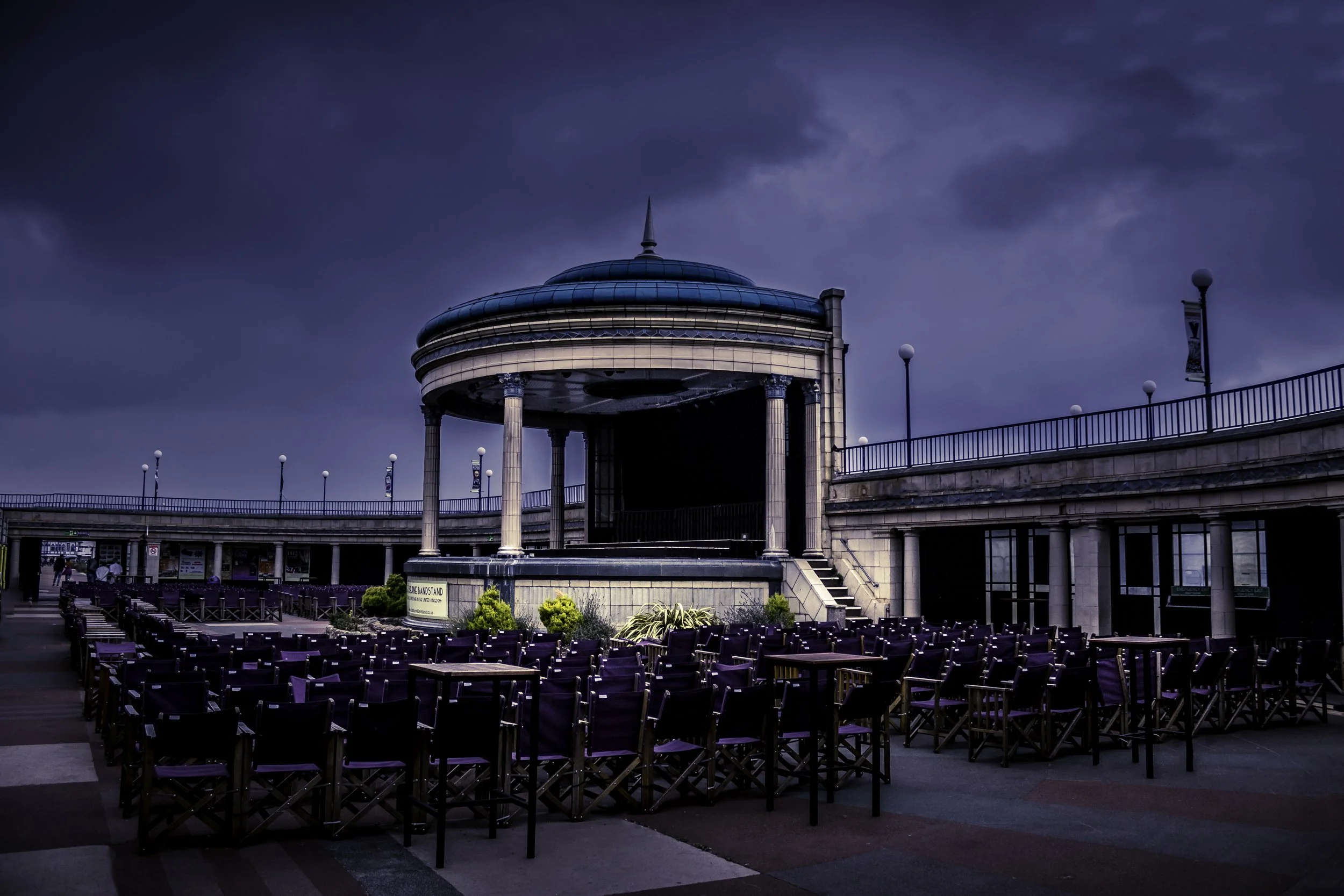 Empty outdoor amphitheater with a circular stage and purple chairs, under a cloudy sky.