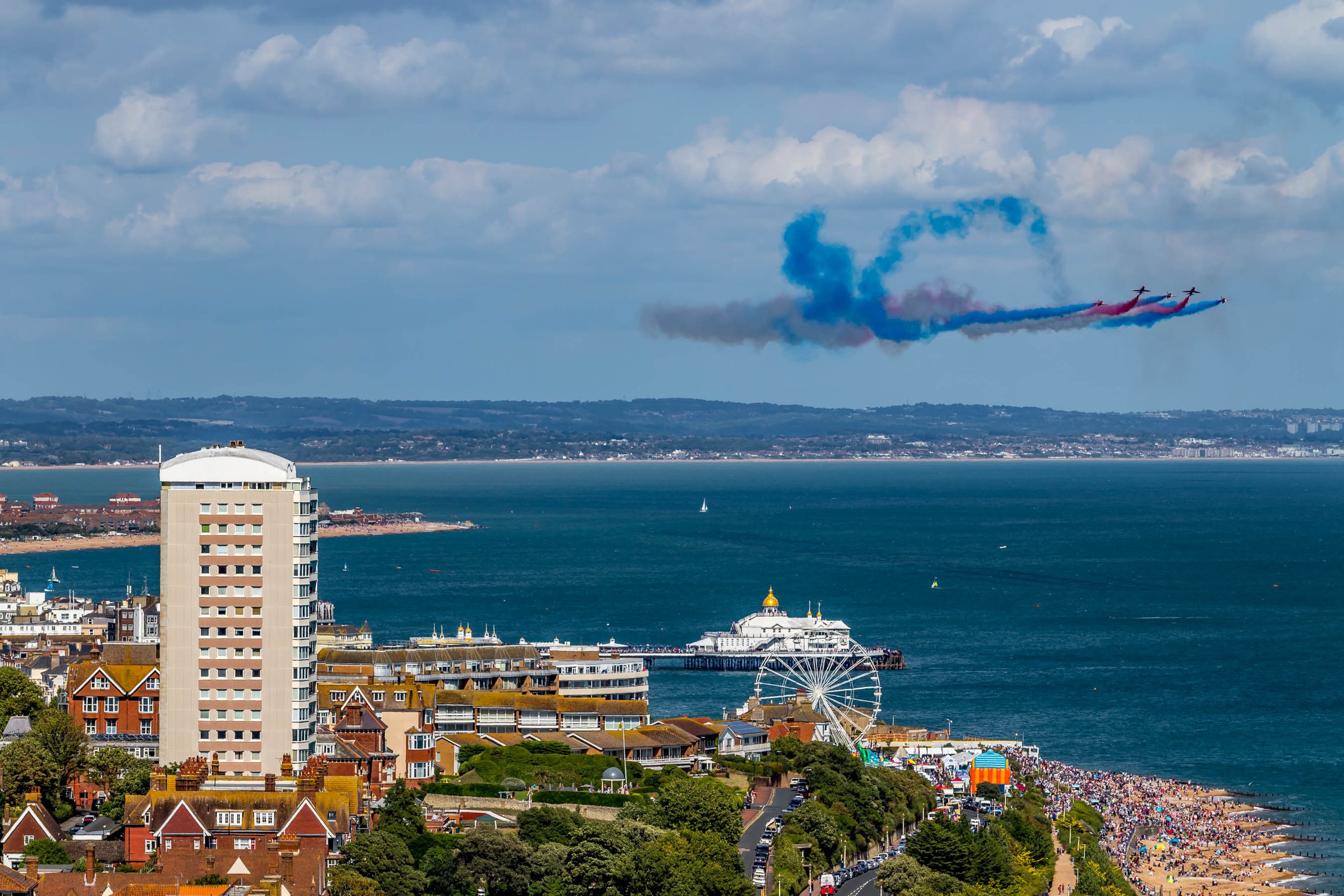 A coastal city with a white high-rise building, a Ferris wheel, and a beach filled with people. Five planes are flying in formation overhead, emitting red, white, and blue smoke.