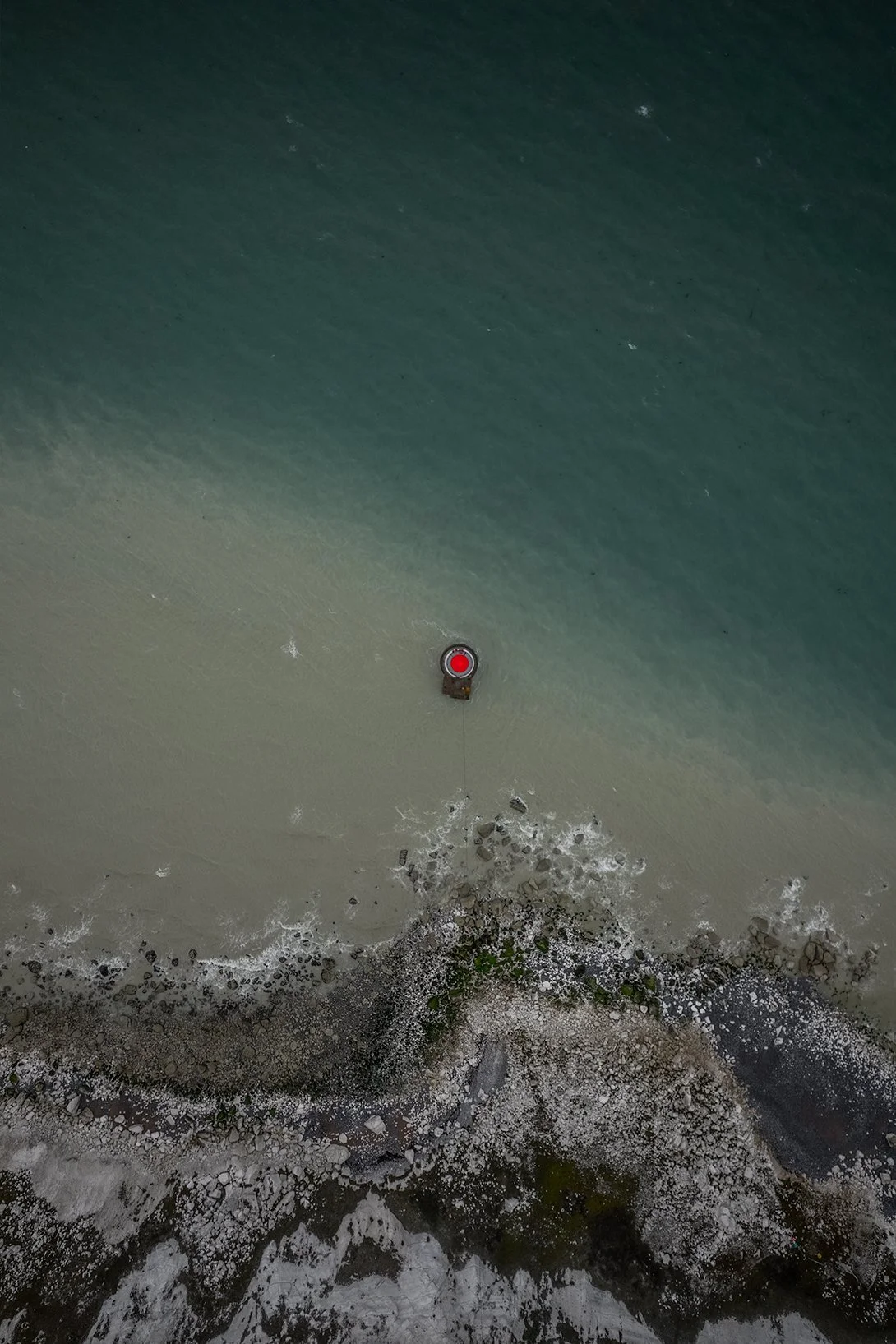 Drone view of a shoreline with a red light and a structure in the water, surrounded by sand, rocks, and small waves.