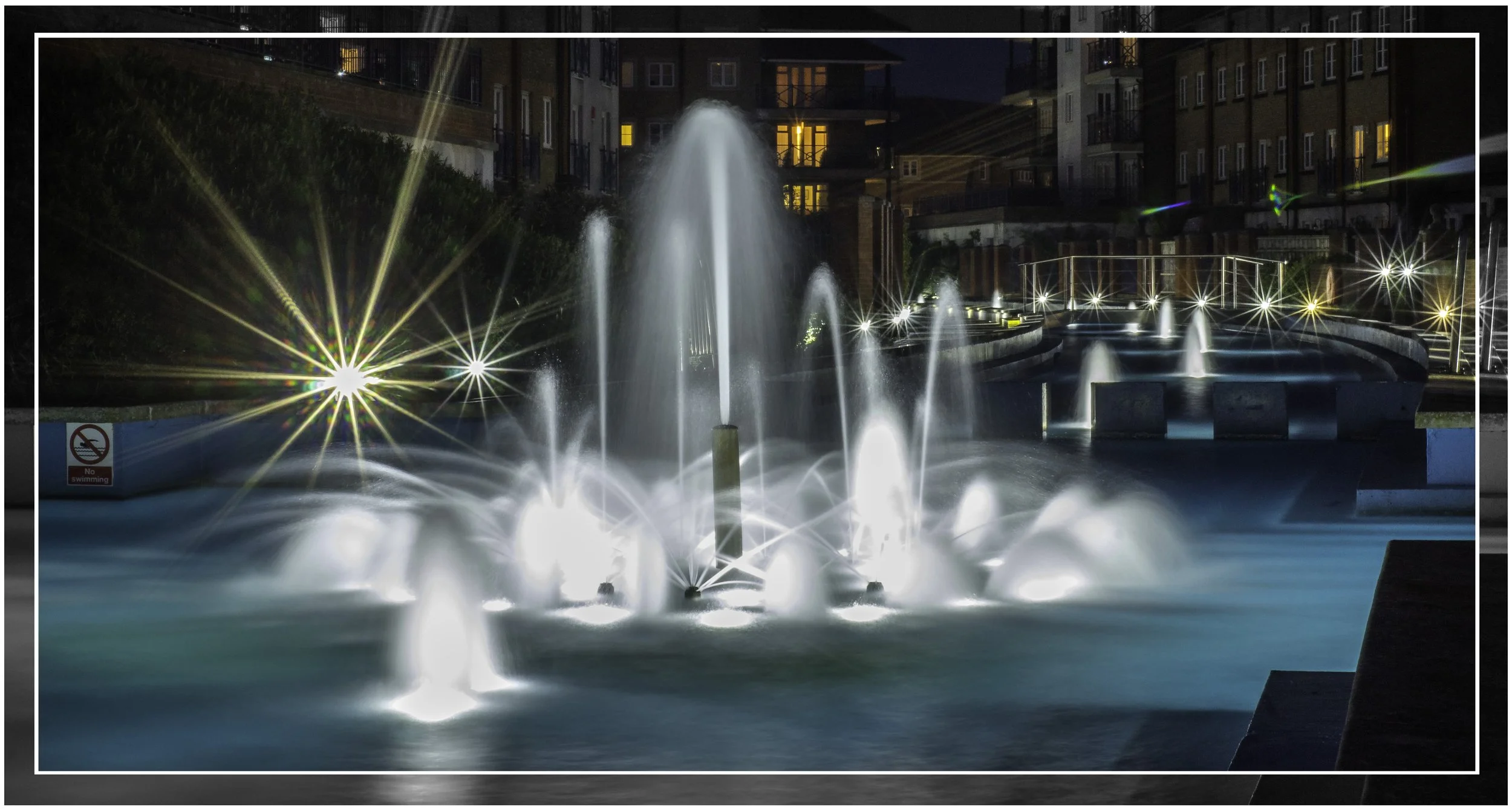 Nighttime view of illuminated fountain with water jets and colorful lights in an urban park, surrounded by residential buildings with lit windows.