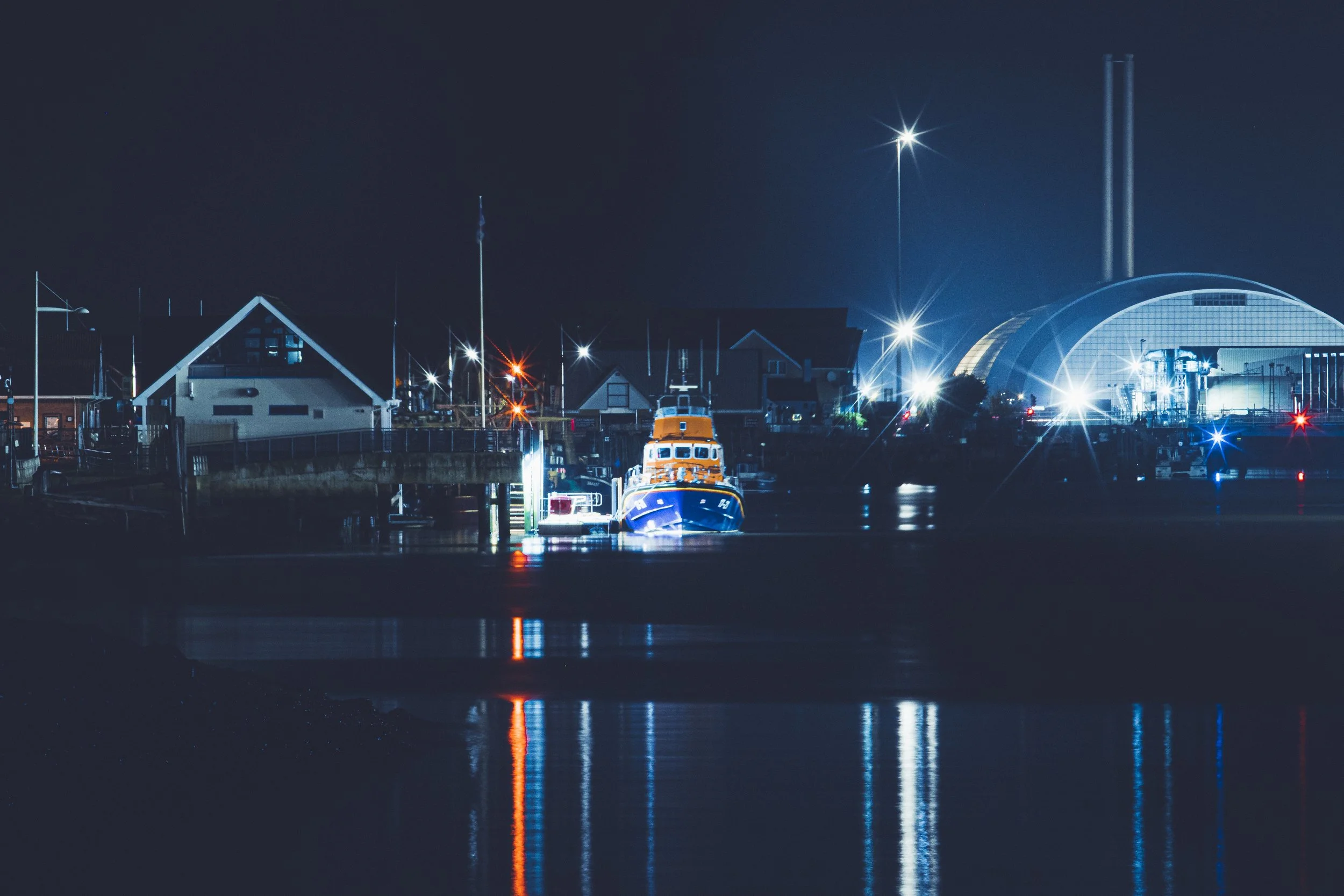 Nighttime scene of a harbor with boats, buildings, and bright streetlights reflecting on the water, featuring a large arched structure on the right.