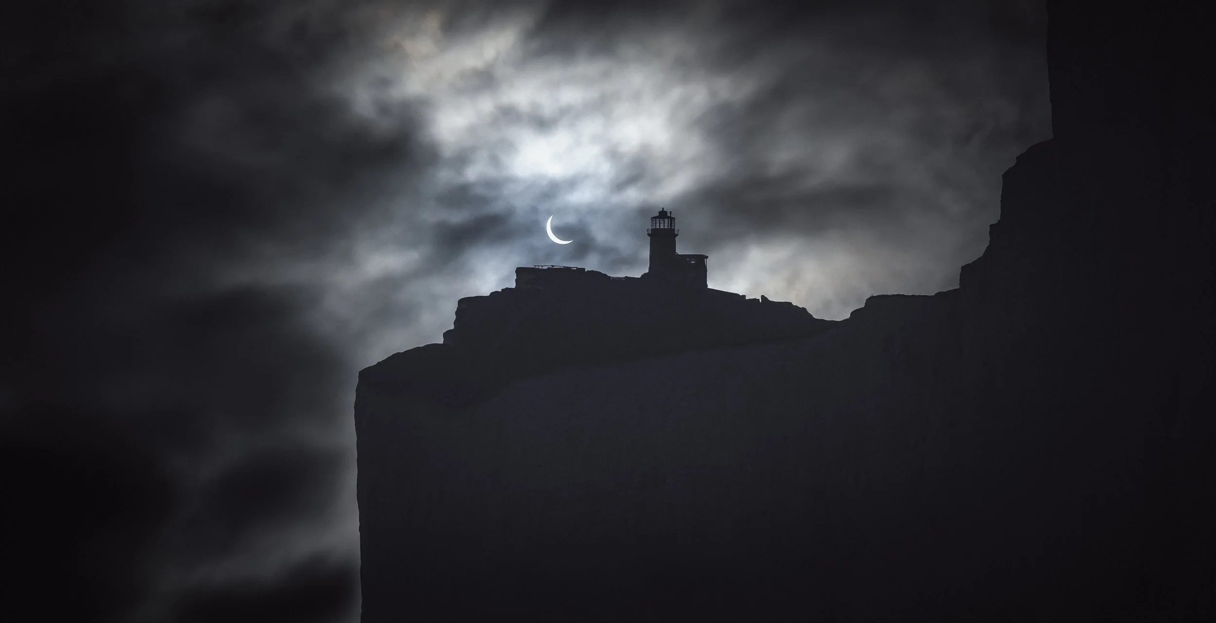 Lighthouse silhouette on a cliff with cloudy night sky and a crescent moon