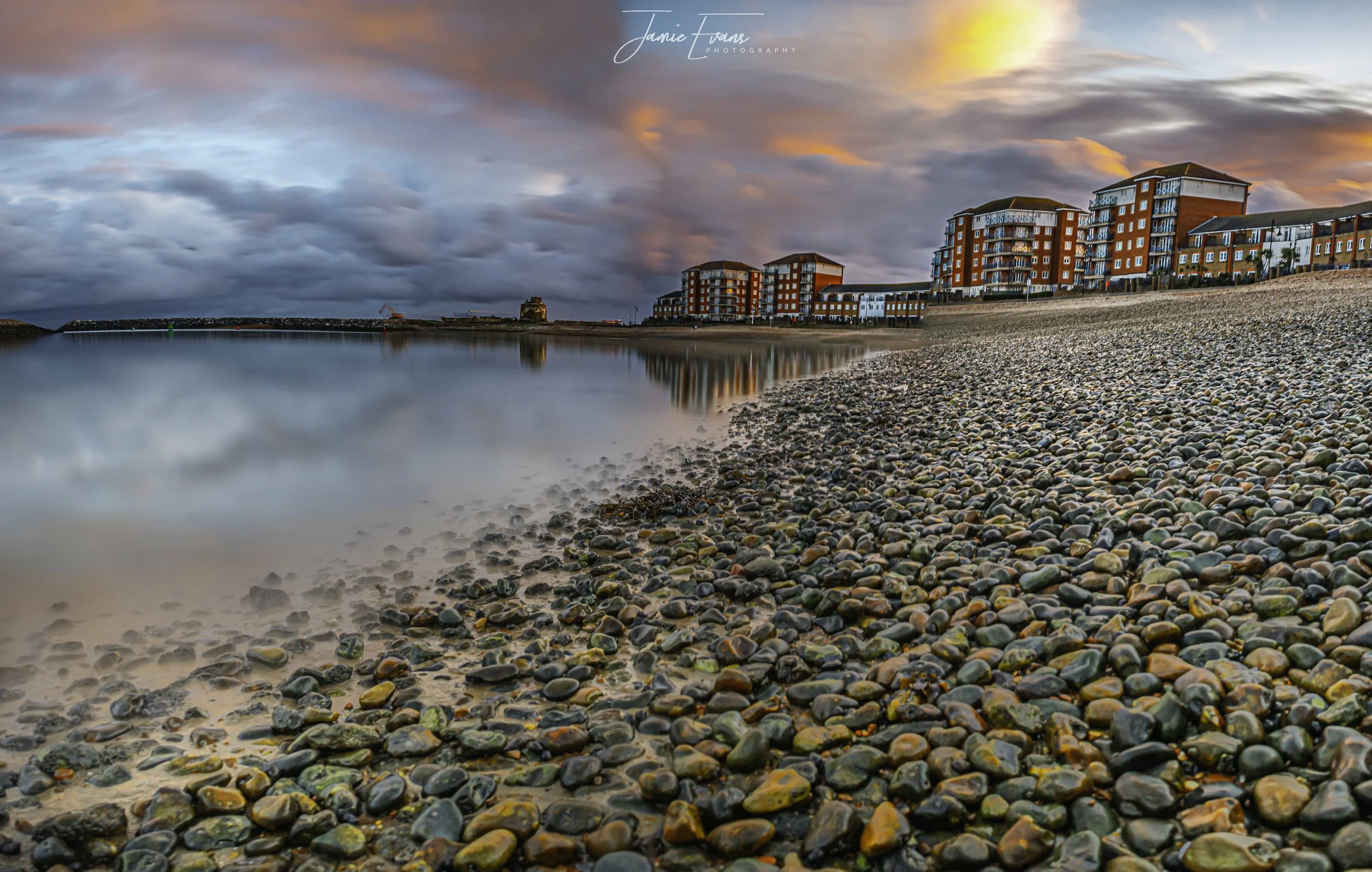 A pebble beach at sunset with a row of modern red and white apartment buildings along the shoreline, and a cloudy colorful sky reflected in the water.