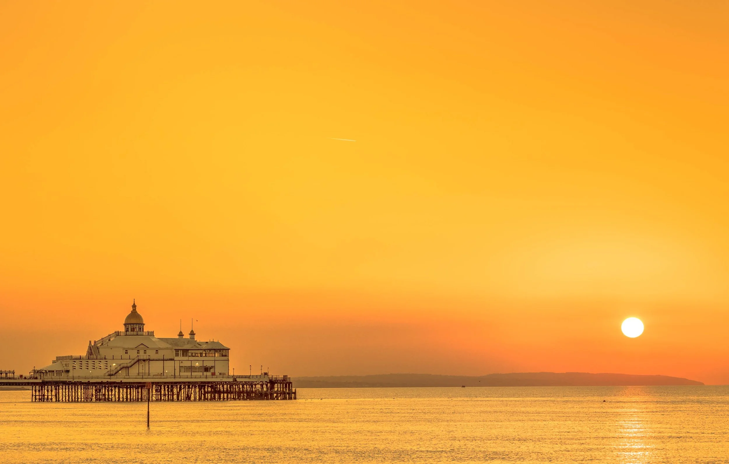 Sunset over the ocean with a pier extending into the water, and a building with a domed roof at the end of the pier. The sky is orange and yellow, with the sun near the horizon.