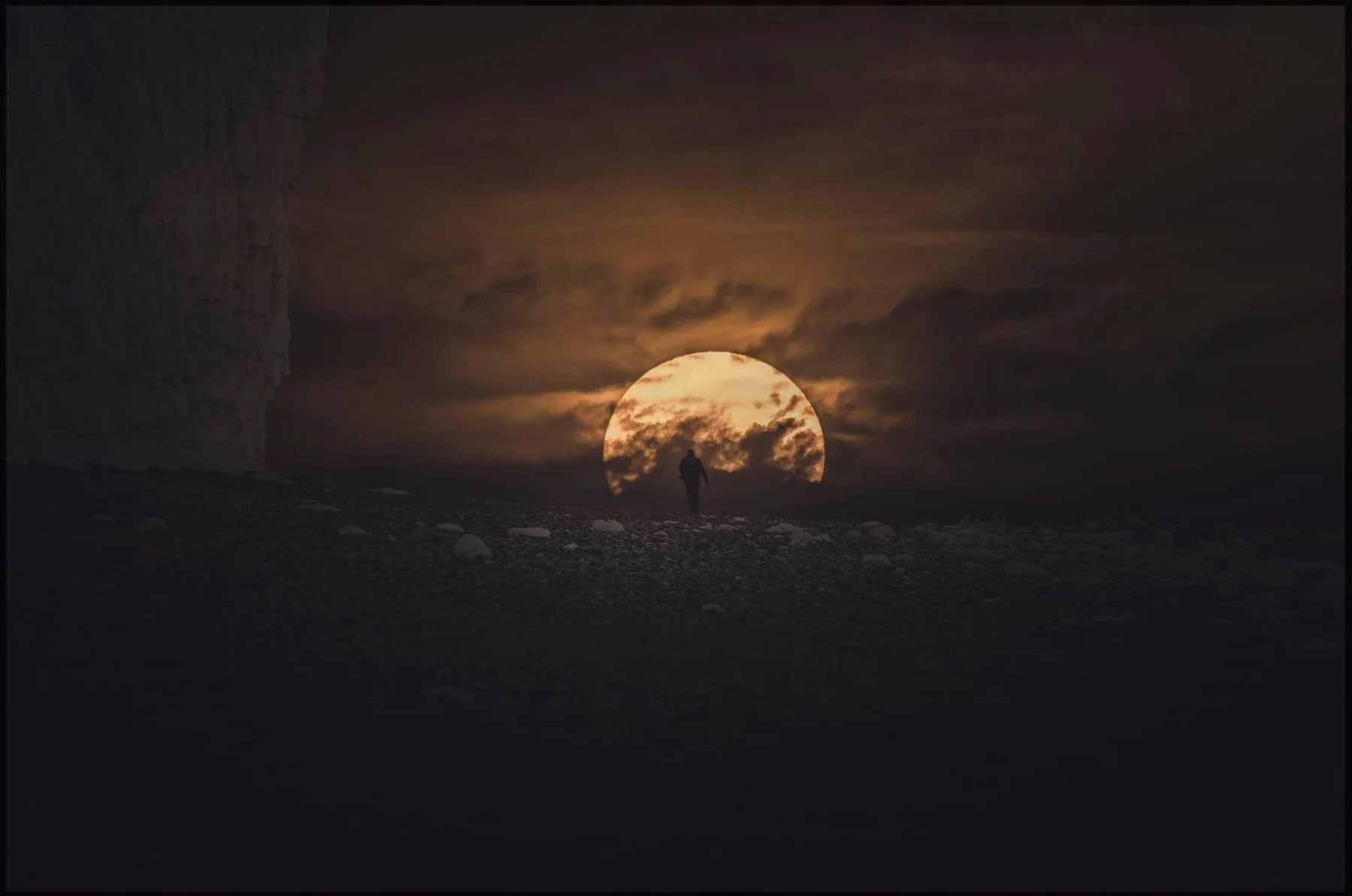 A person walking towards a large, glowing moon partially covered by clouds, with a dark, rocky landscape and a cliff on the left.