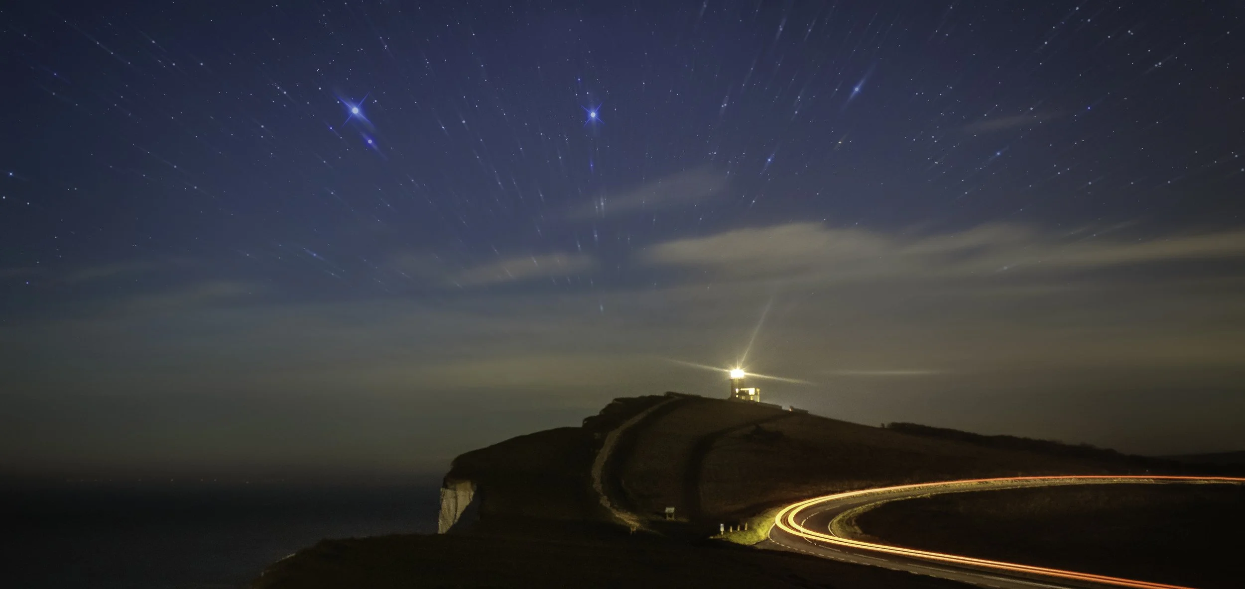Nighttime landscape showing a lighthouse on a hill with star trails in the sky and light trails from moving vehicles on a curving road.