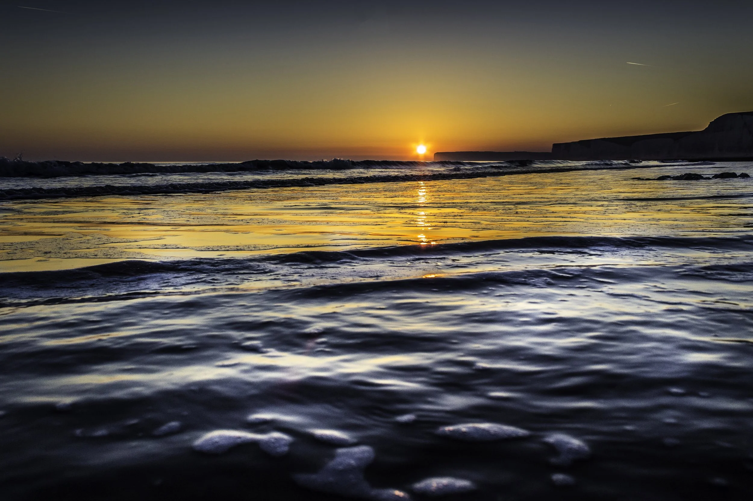 Sunset over the ocean with waves and rim of cliffs in the distance.