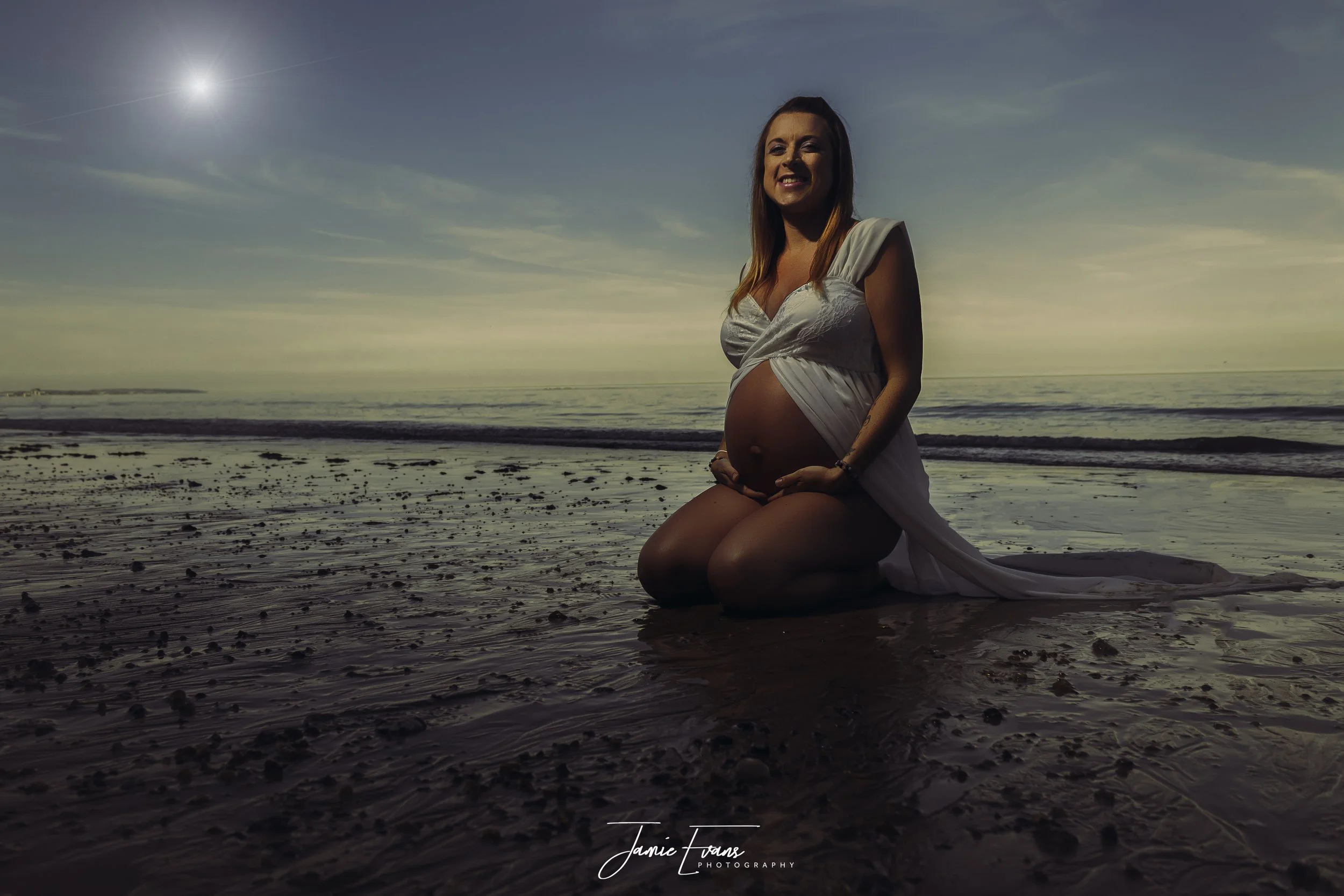 Pregnant woman kneeling on the beach at sunset, wearing a white dress, with the ocean and sky in the background.