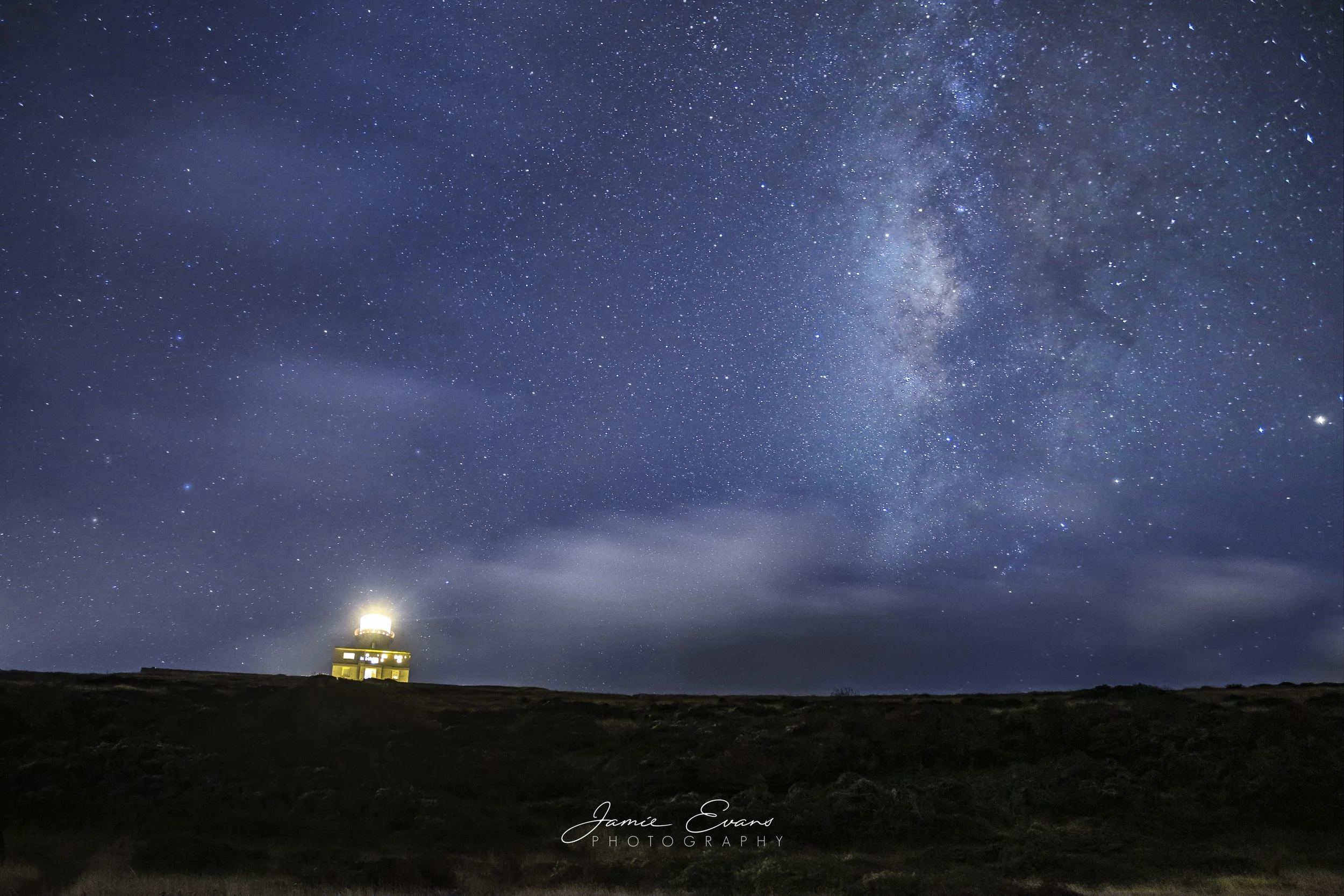Night sky filled with stars and the Milky Way galaxy above a lighthouse on a dark landscape.