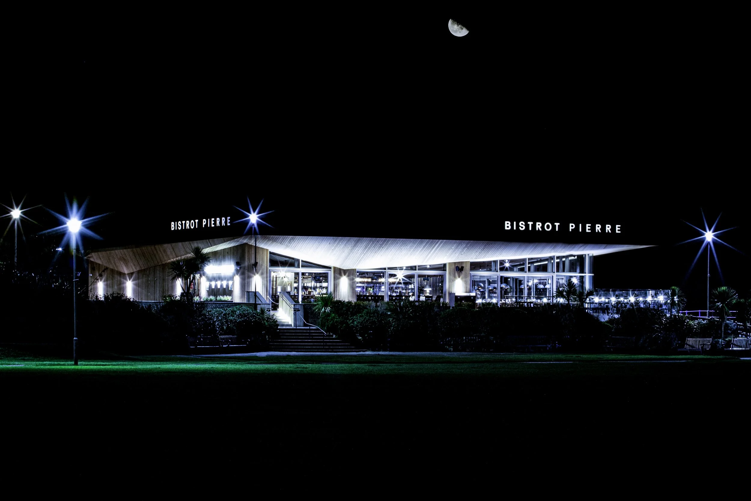 Night view of Bistrot Pierre restaurant with illuminated sign, glass windows, and surrounding greenery under a dark sky with a visible moon.