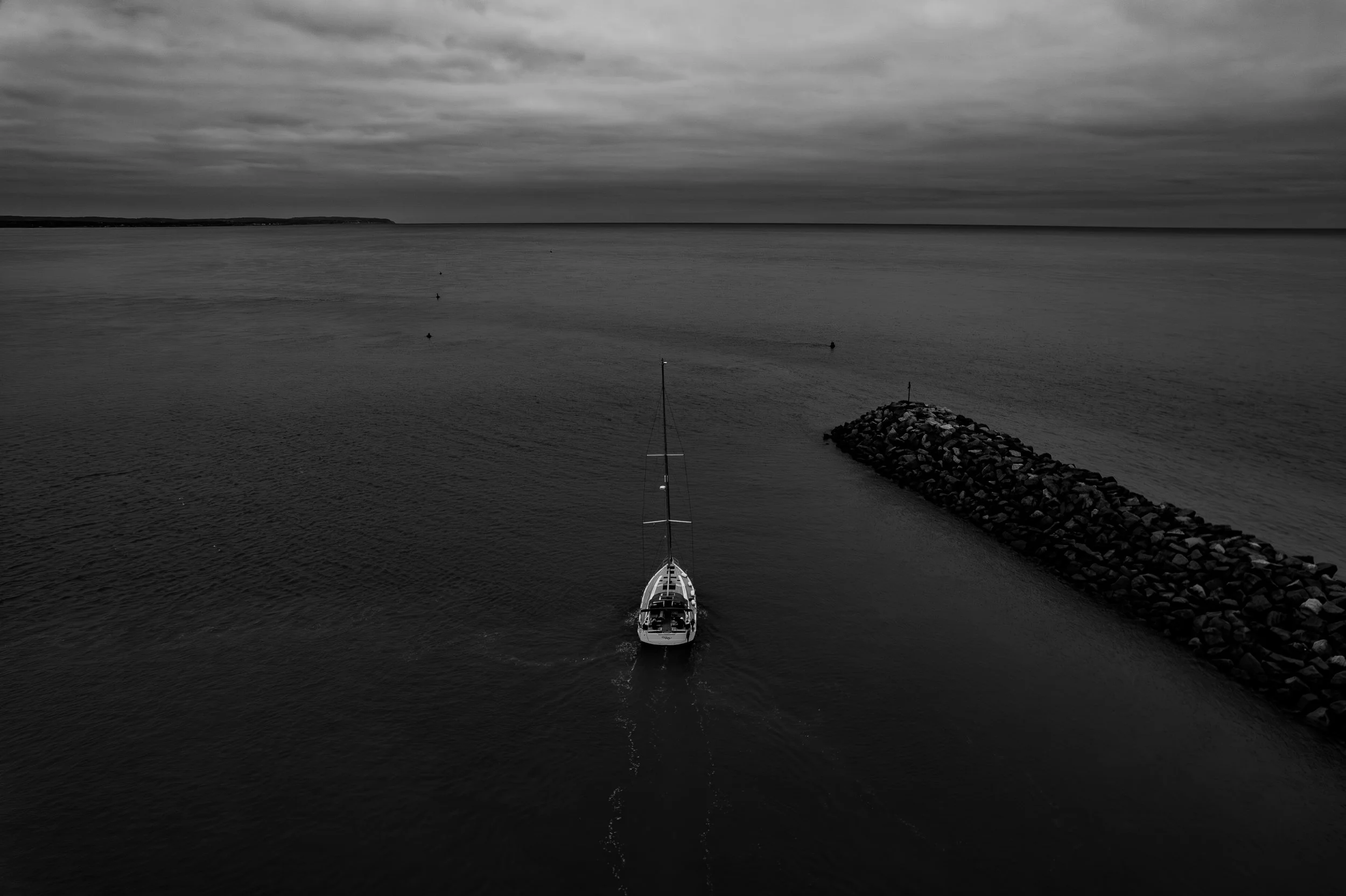 A sailboat sailing near a rocky breakwater in a calm body of water under cloudy skies, in black and white.