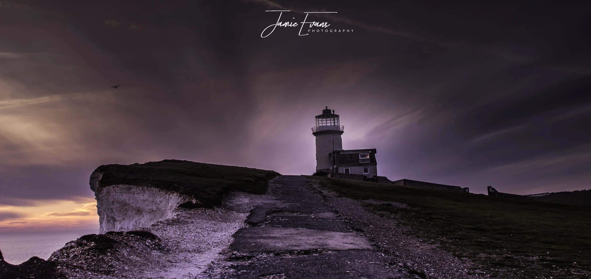 Lighthouse on a hilltop during sunset with dark clouds in the sky.