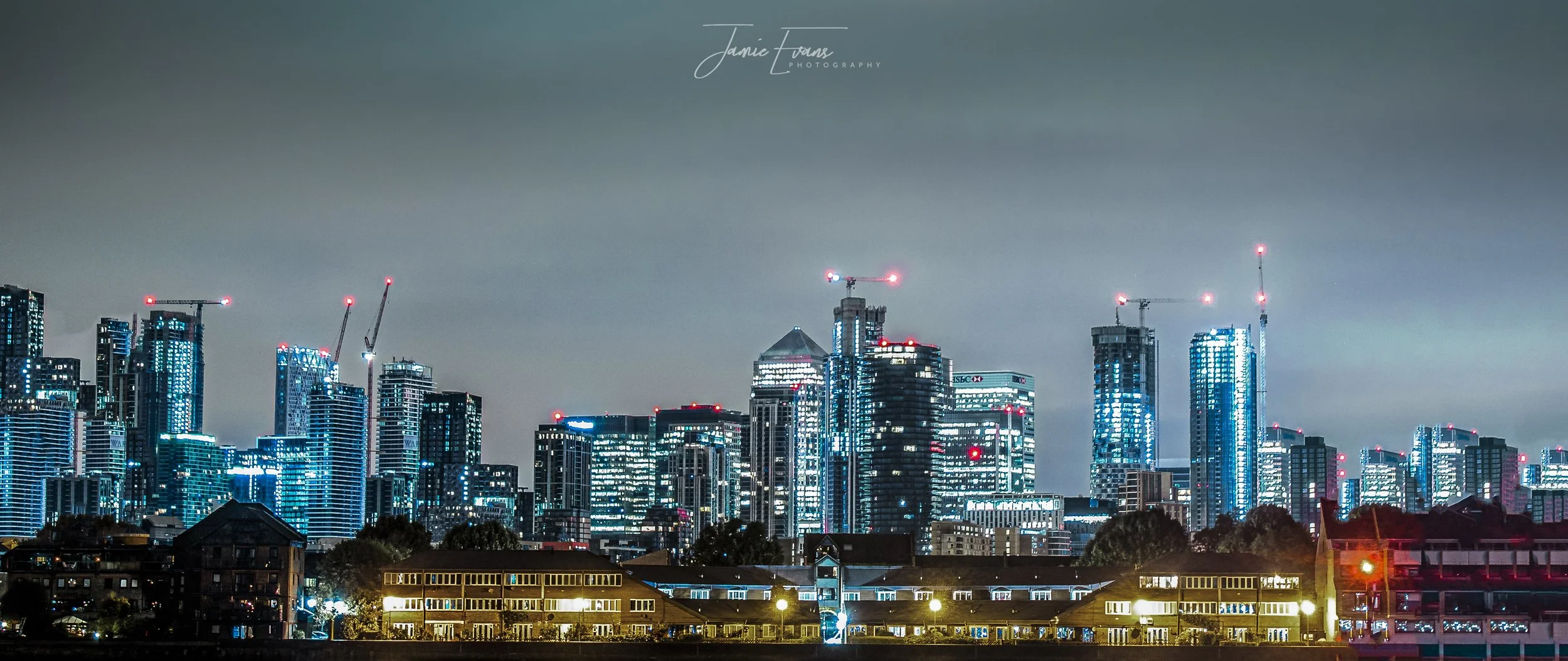 Nighttime city skyline with illuminated tall skyscrapers under a cloudy sky, showing ongoing construction cranes with red lights on top.