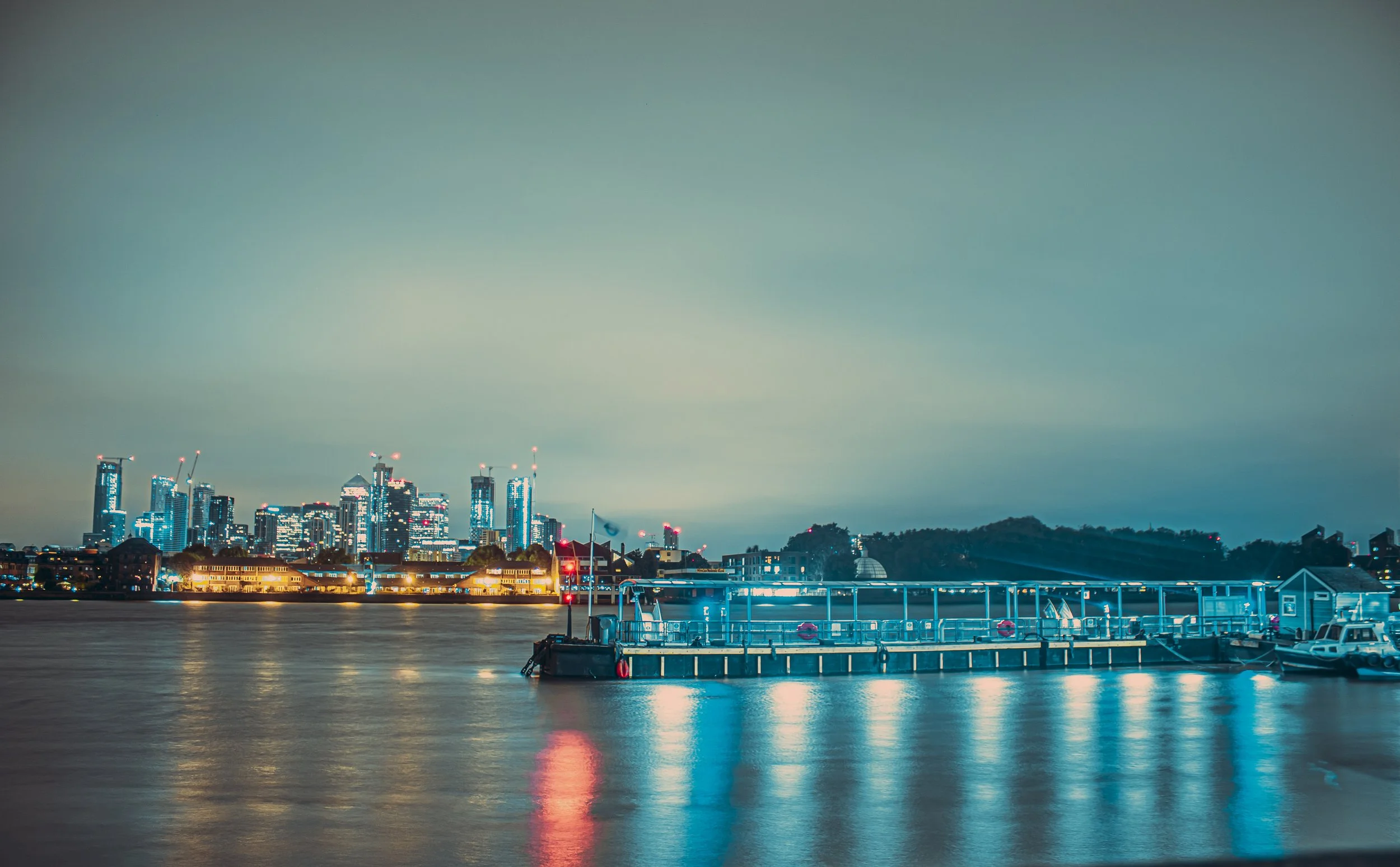 Night view of a city skyline with tall buildings under construction across a body of water, featuring a docked boat and calm reflections on the water.