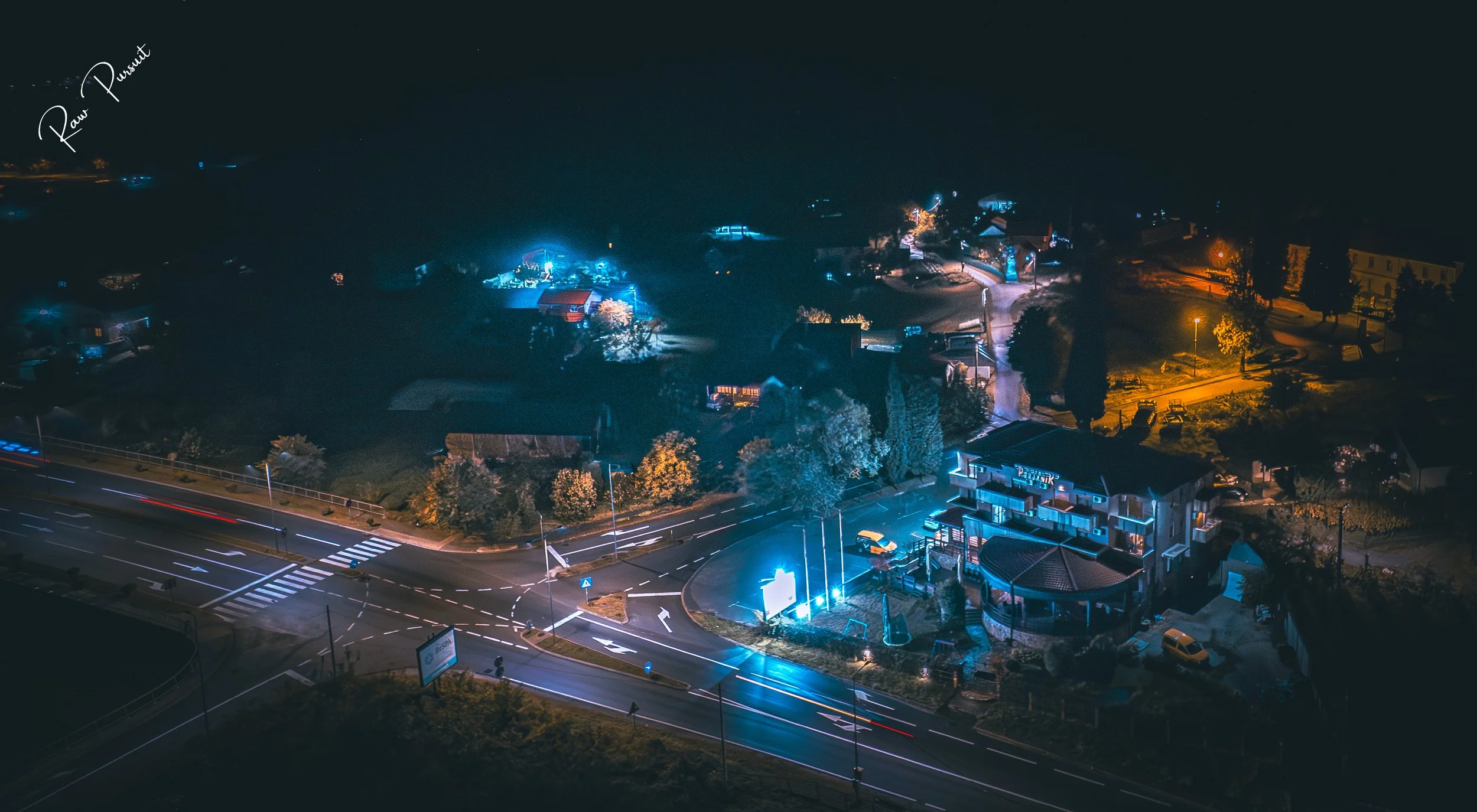 Nighttime aerial view of a small town with illuminated buildings, streetlights, and roads, featuring a prominent building with bright blue neon lights on the right side of the image.