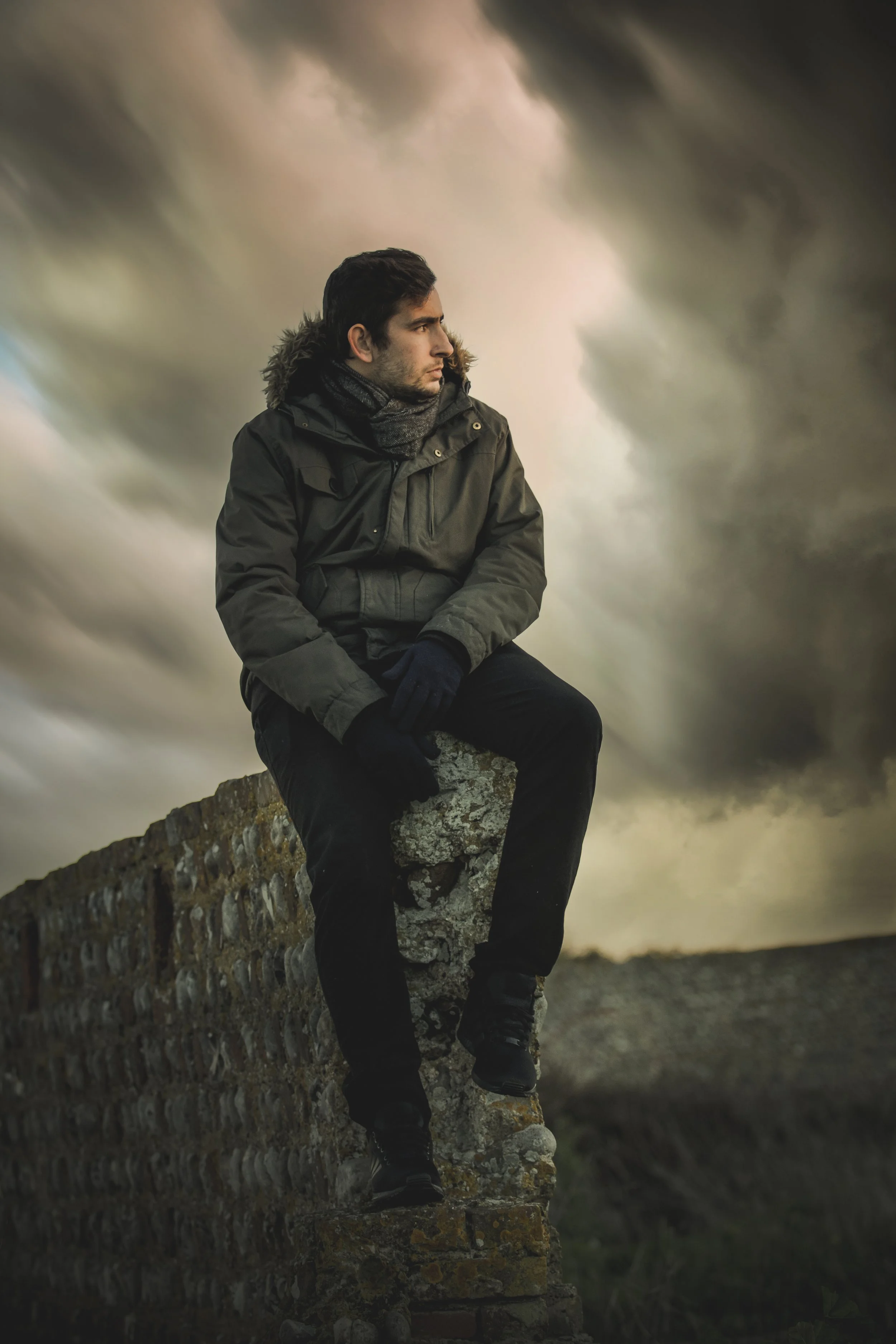 A man sitting on a stone wall outdoors during cloudy weather, looking to his left, dressed in a winter jacket and gloves.