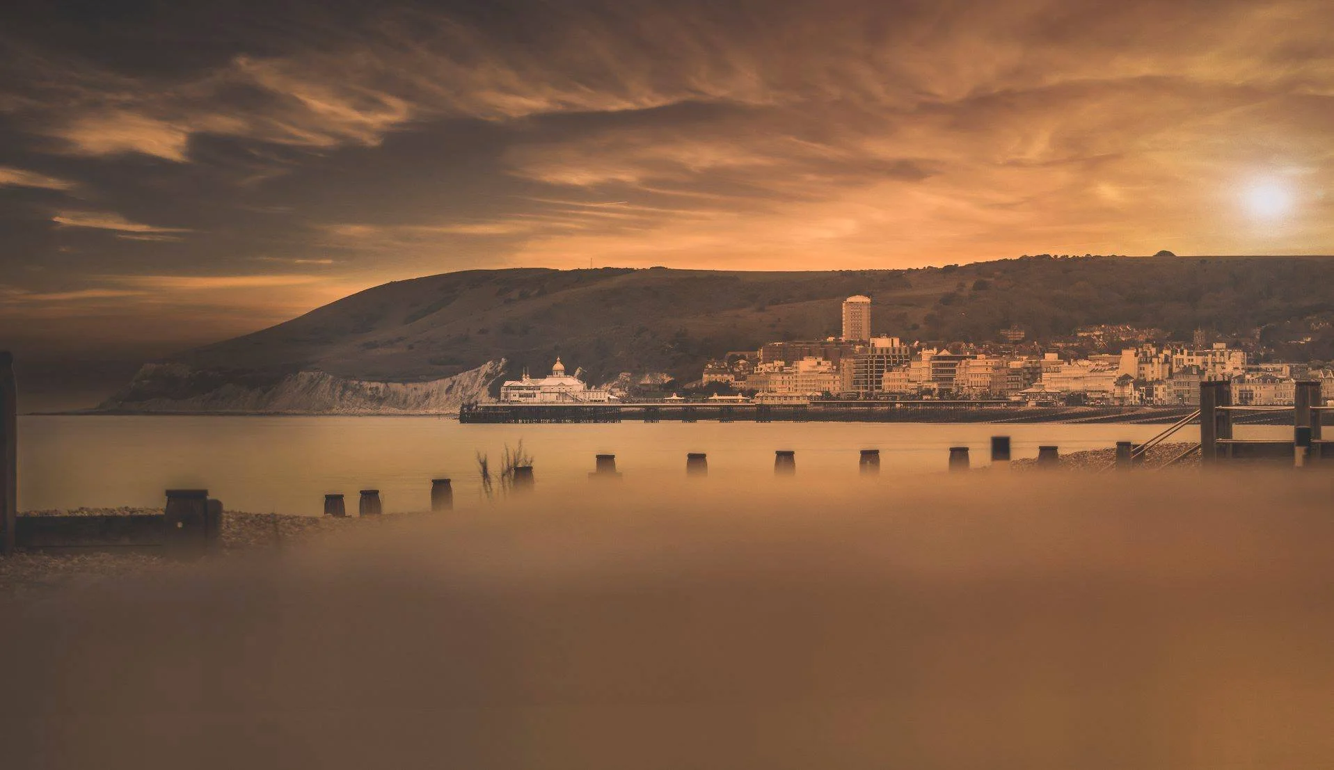 A calm waterfront view at sunset with a city skyline and hills in the background, featuring several structures along the shore and a partially submerged dock.