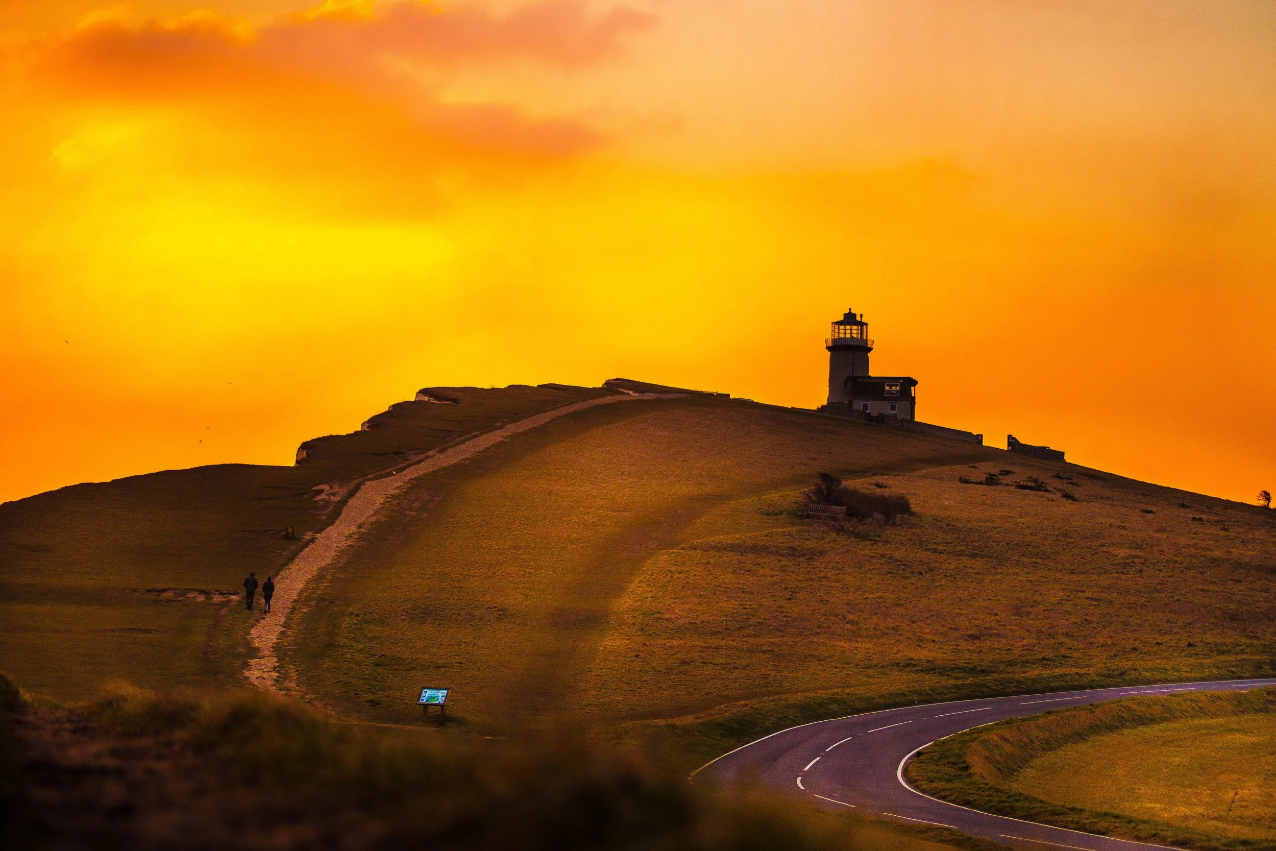 A lighthouse on a hill at sunset with a winding road and two people walking along a path.