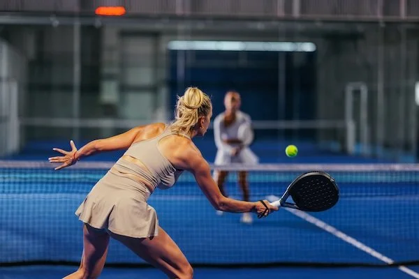 A woman playing pickleball on an indoor court with a man standing nearby in the background.