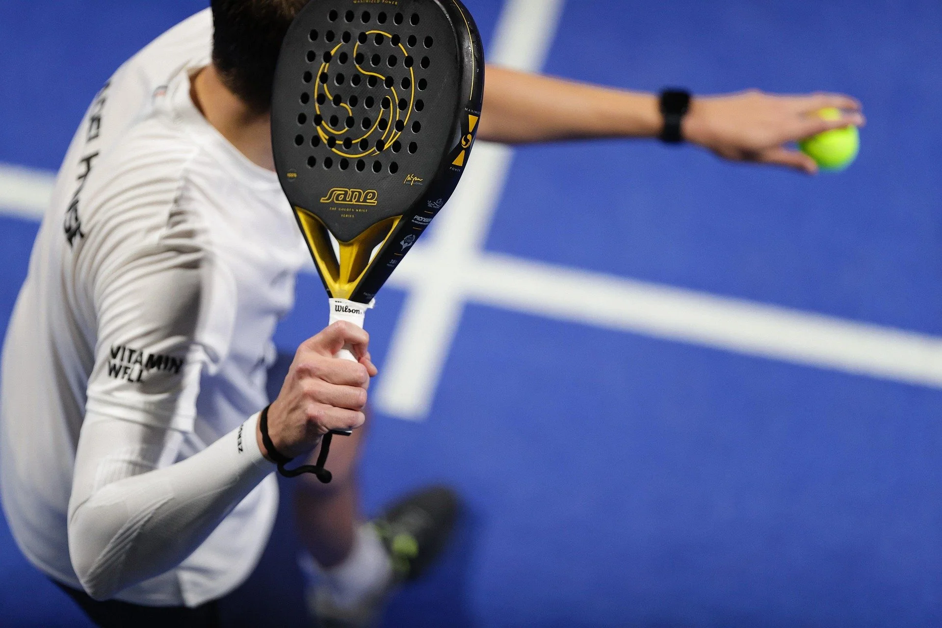 A tennis player is about to serve, holding a tennis racket in front of their face and a tennis ball in the other hand, on a blue court.