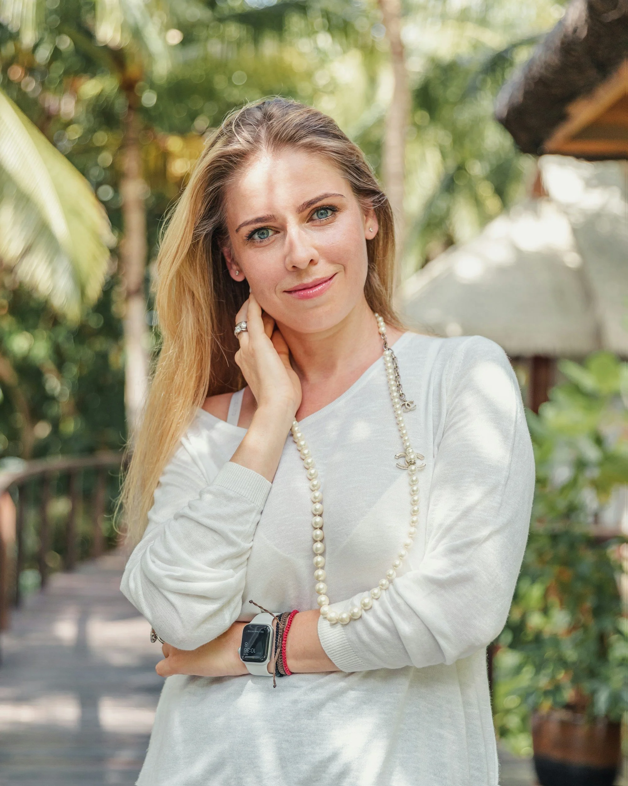 A woman with long blonde hair and blue eyes stands outdoors in a lush, green environment, wearing a white long-sleeve top, a pearl necklace, and a smartwatch on her wrist, smiling softly and touching her neck with one hand.