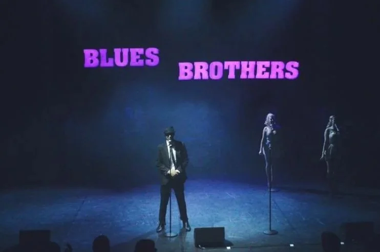 Stage with three performers, one man in a suit and hat at microphone, two women in dresses with microphones, and a sign overhead reading 'Blues Brothers' in purple letters.